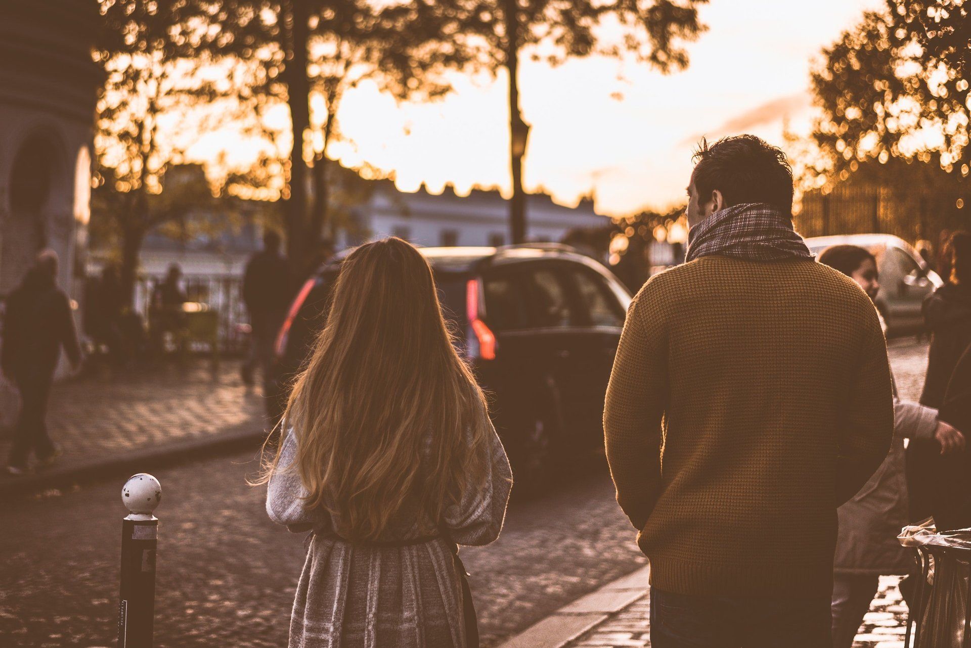 Two people walking down a wet city street at sunset, with warm lights and cars in the background