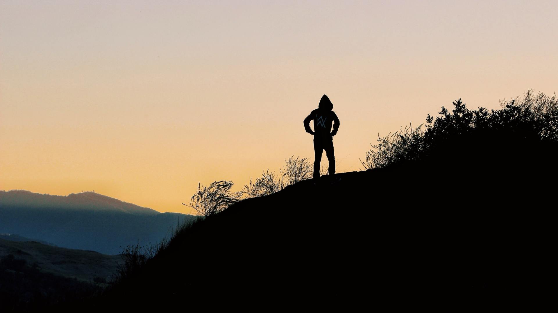 Silhouette of person atop a hill at sunset with hands on hips, overlooking mountain range.