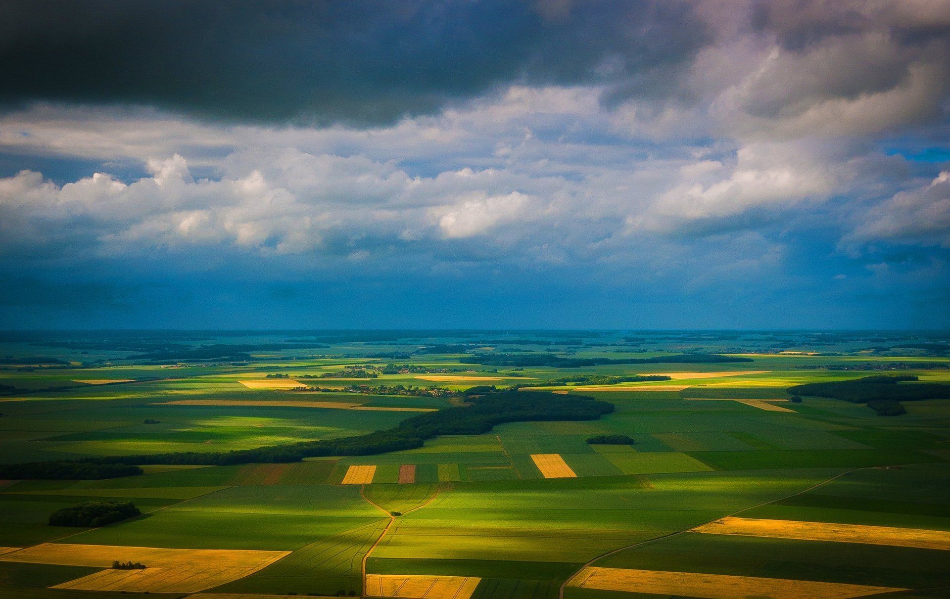 An aerial view of a patchwork of green and gold fields under a vast, dramatic, cloudy sky.
