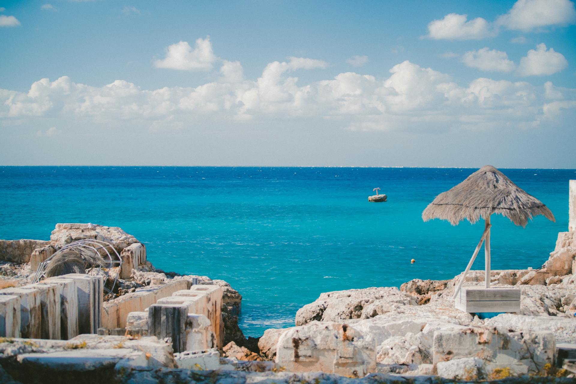 Bright blue ocean scene with small boat, thatched roof umbrella, and rocky shoreline under a partly cloudy sky.