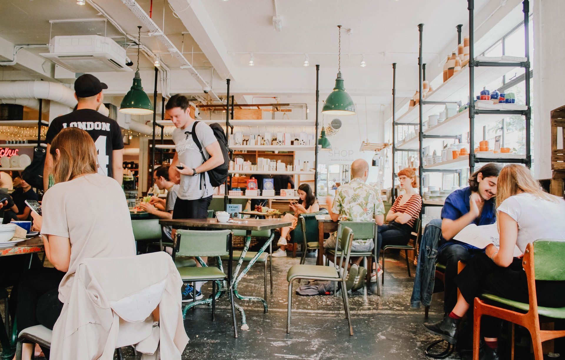 Een druk caféinterieur met mensen die zitten, praten en werken aan tafels onder hangende lampen.