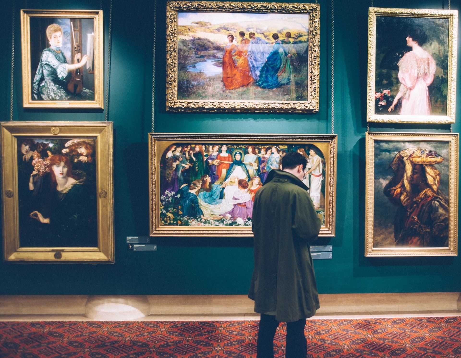 Person in green coat views paintings in an art gallery; gold frames on teal wall.