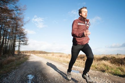 Man running on a dirt path in a rural area, wearing a maroon jacket and black pants.