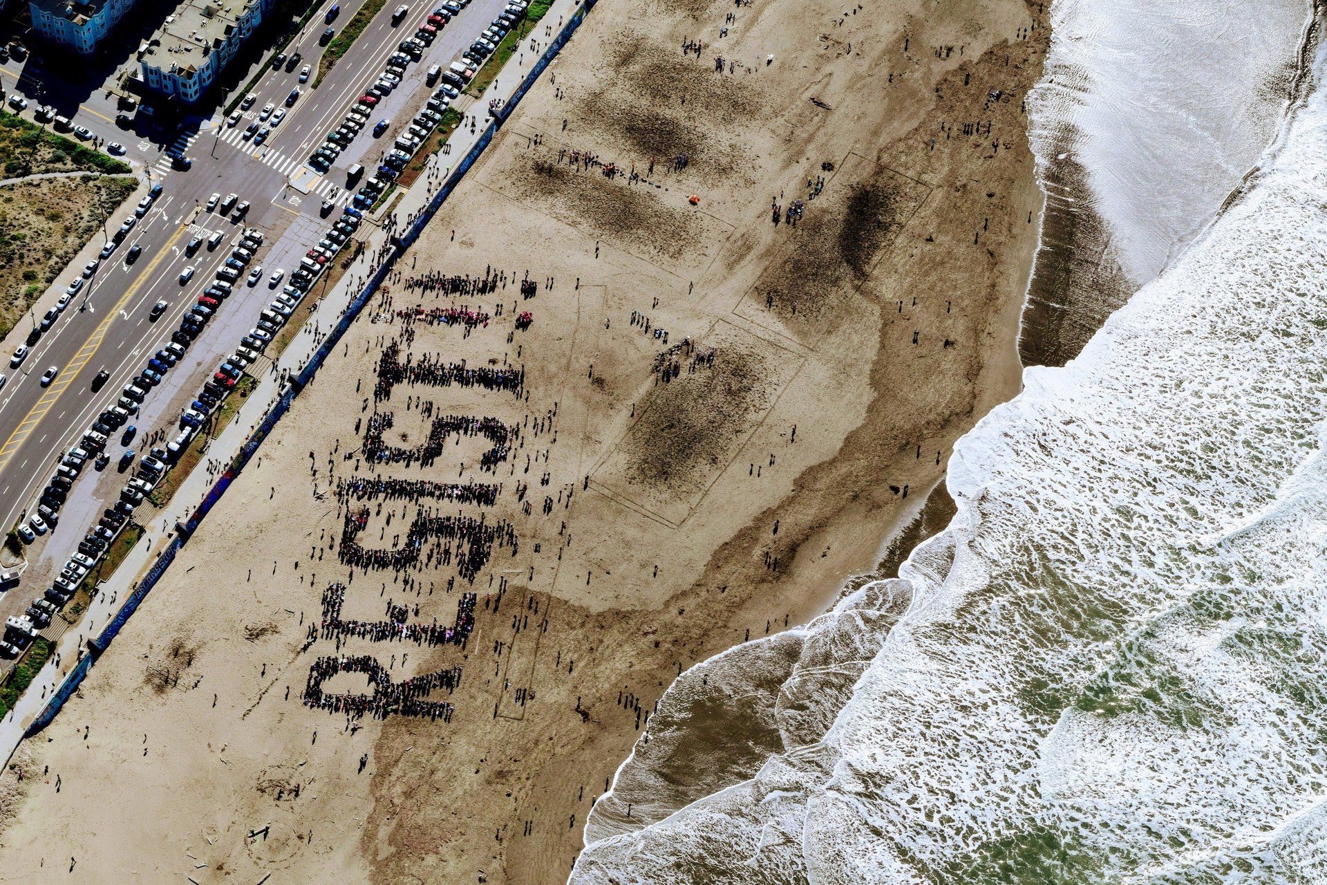 A large group of people stand on a sandy beach to form the word