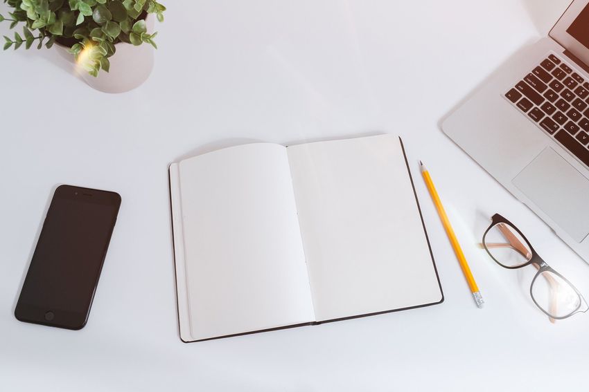 White desk with laptop, notebook, pencil, phone, eyeglasses, and plant.
