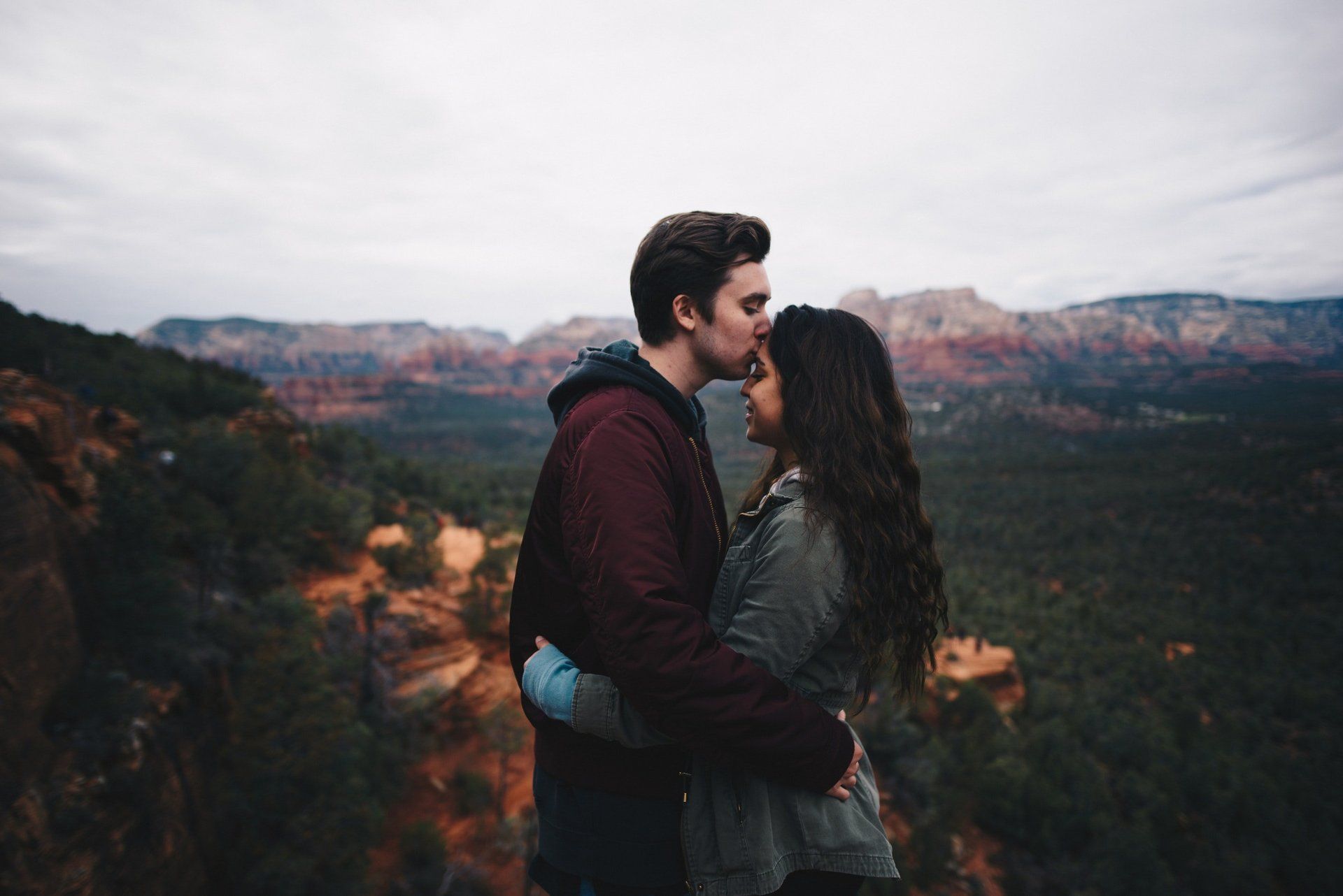 Couple embraces on a cliff with mountainous vista; man kisses woman's forehead, both facing the view, overcast sky.