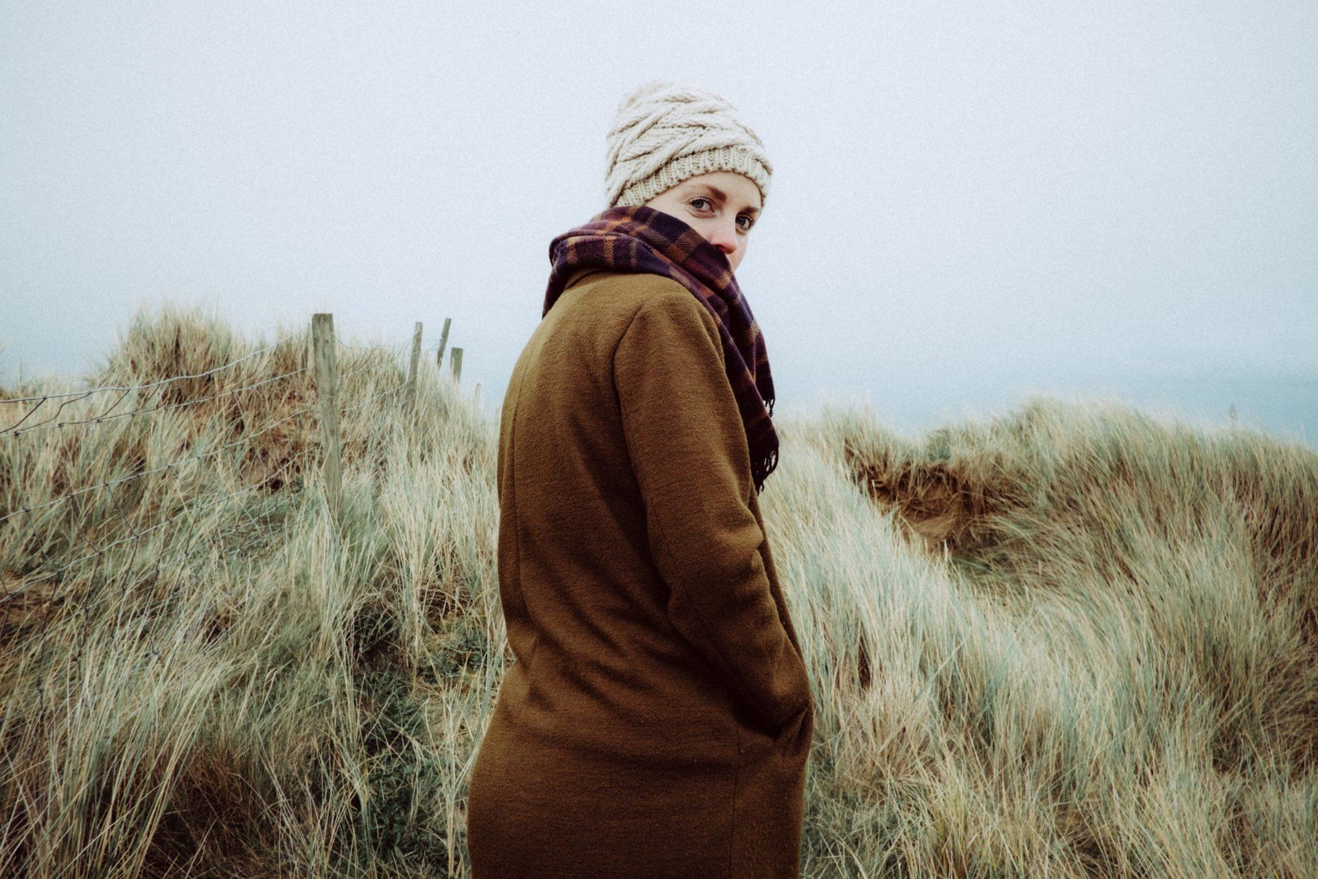 Person in a brown coat and knit hat standing in tall grass under an overcast sky