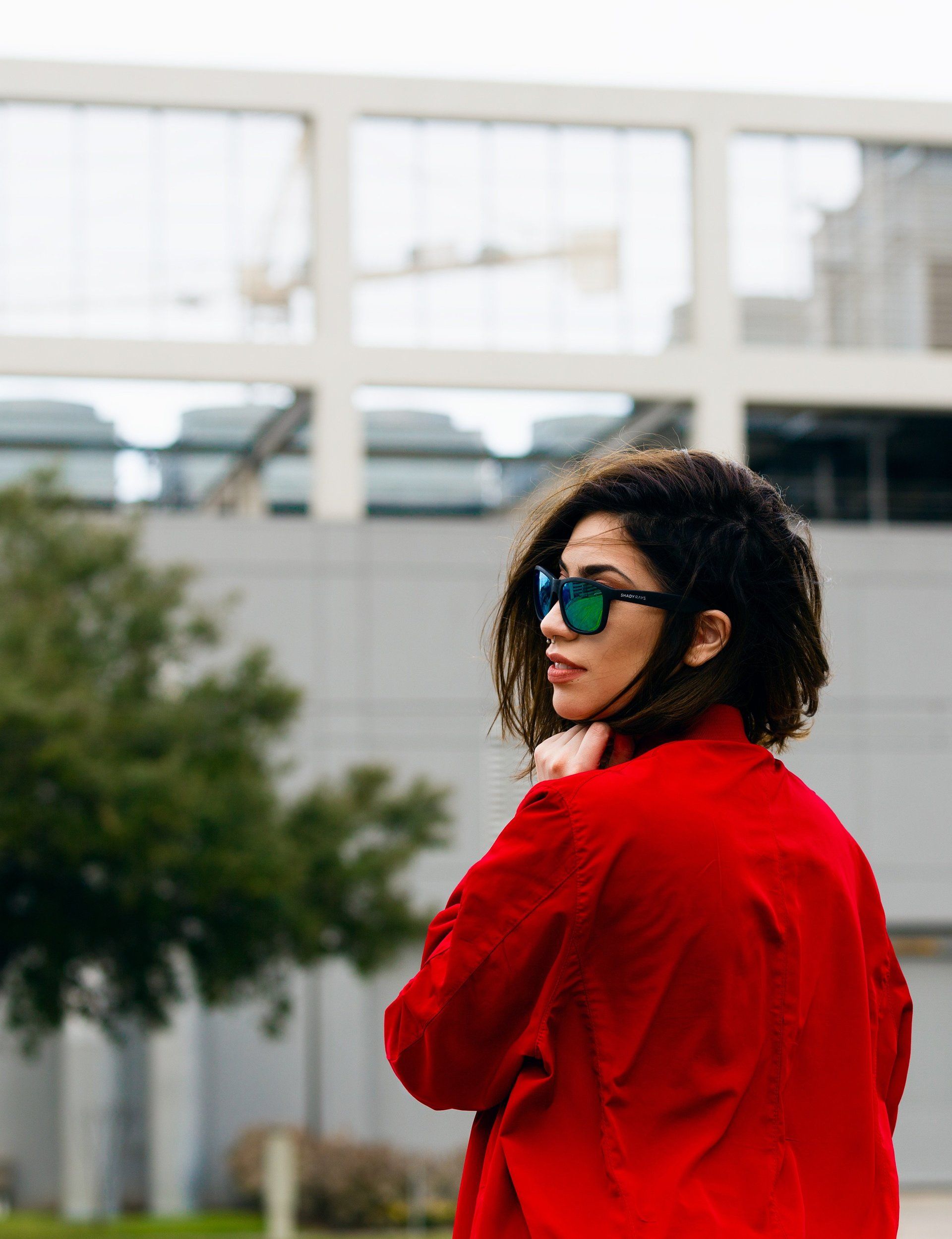A woman wearing sunglasses and a red jacket is standing in front of a building.