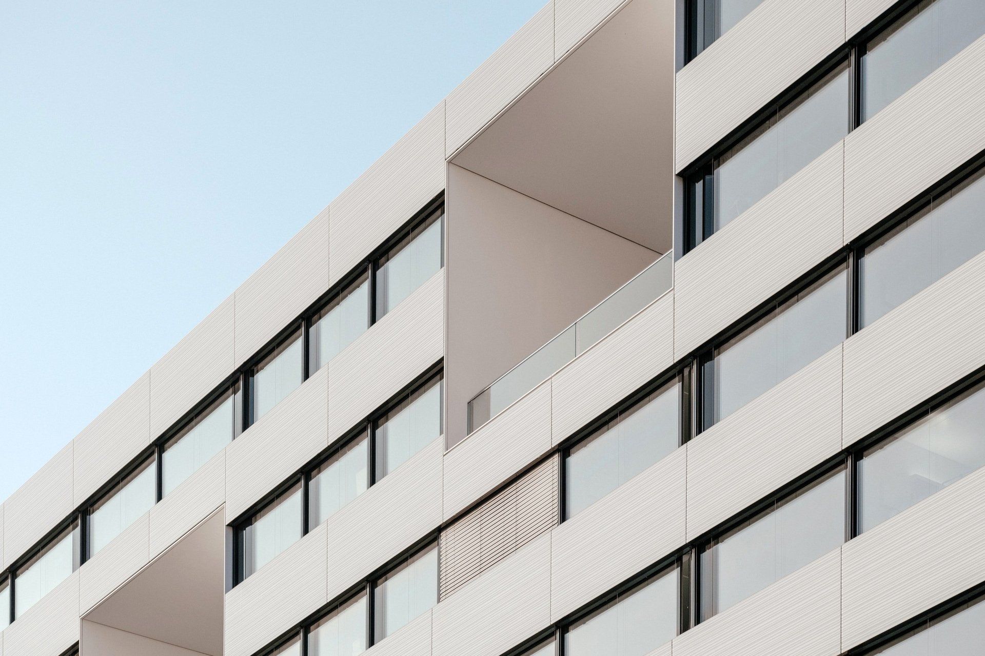 Moderne weiße Gebäudefassade mit quadratischen Balkonen, Fenstern und hellblauem Himmel.
