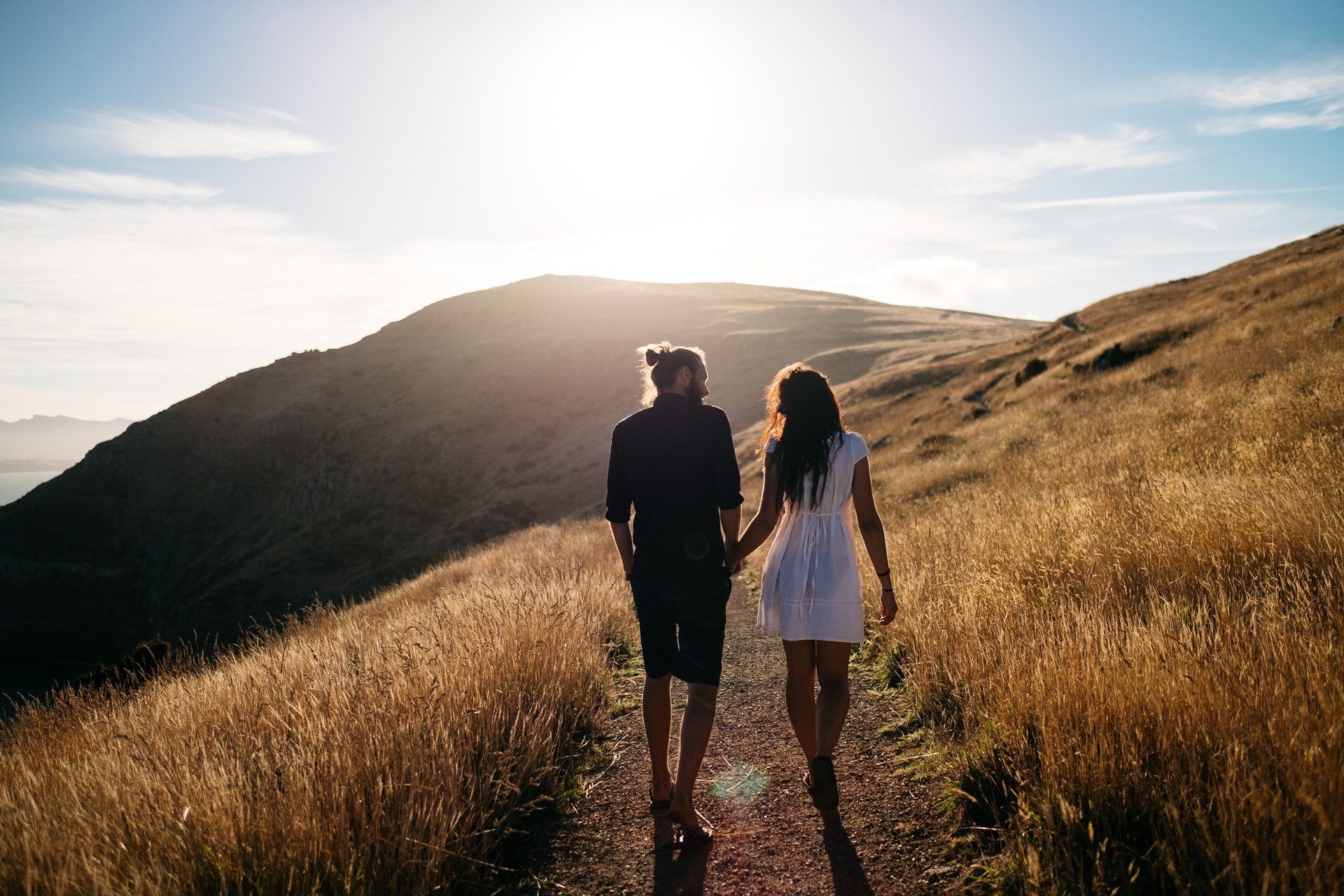 Two people walking on a sunlit grassy hillside at sunset, facing a bright horizon