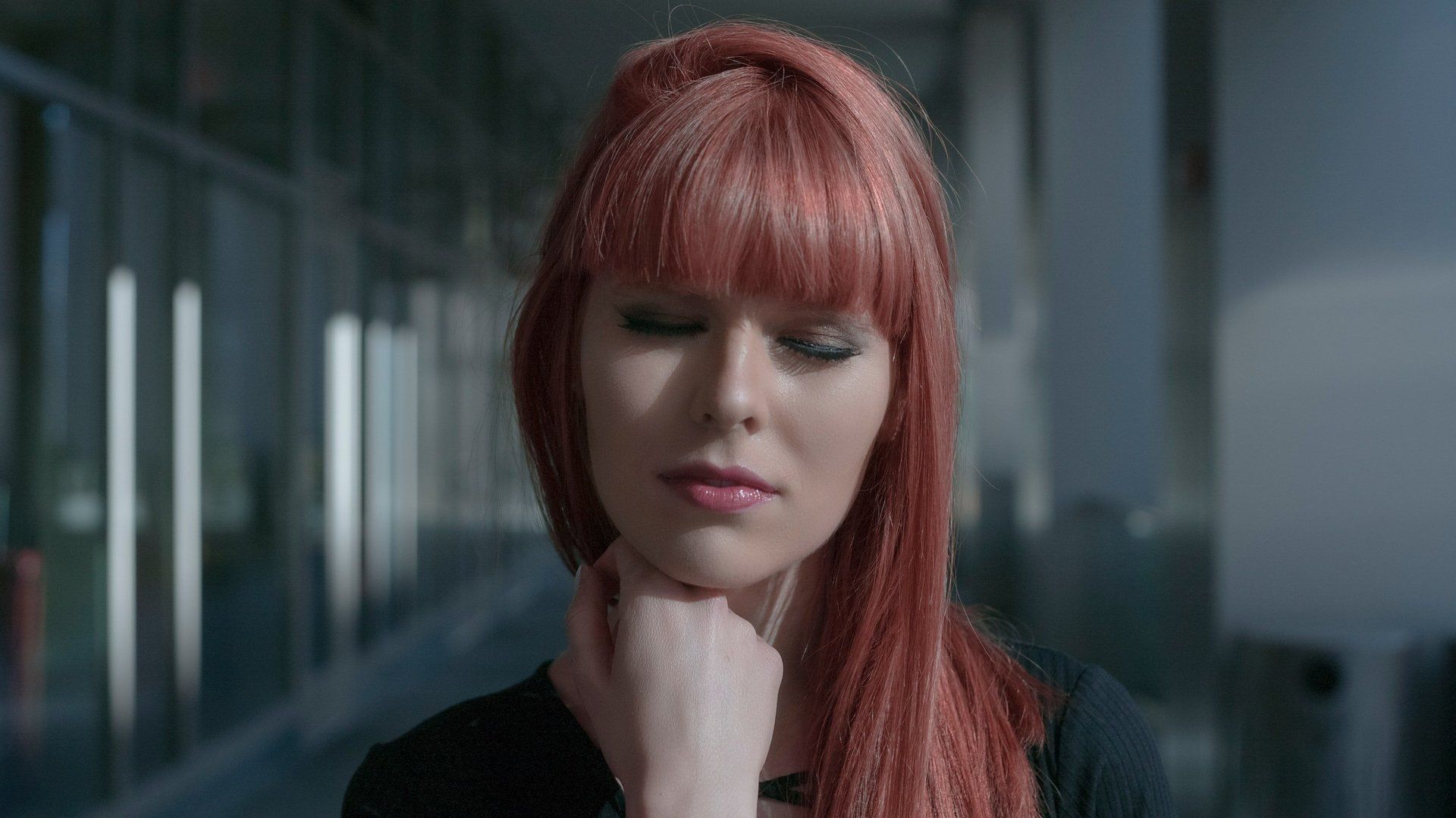 Woman with red hair, hand on chin, looking down, possibly thoughtful, outside a modern building.