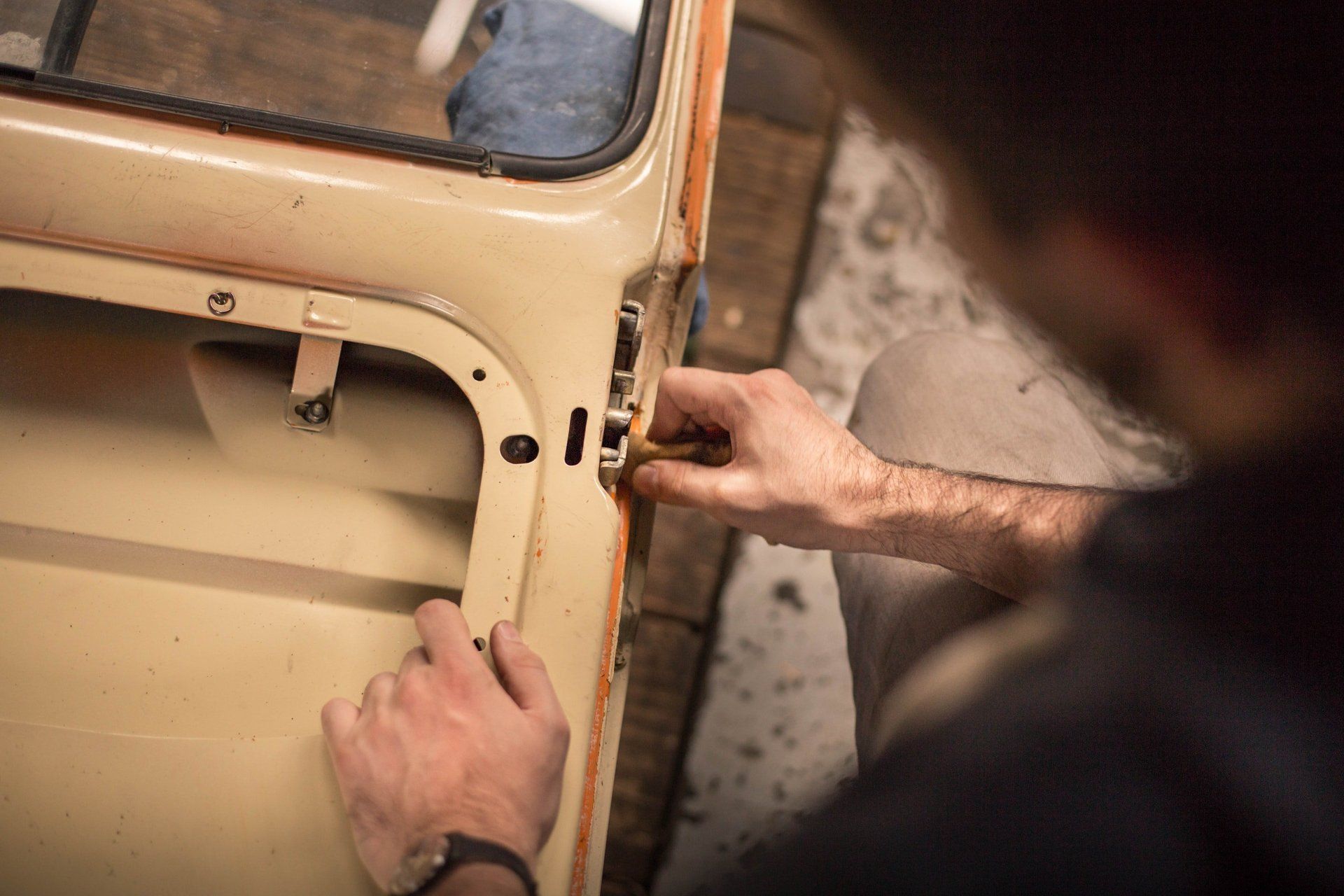 Person working on a car door lock, in a workshop. Hands using a tool on a cream-colored door.
