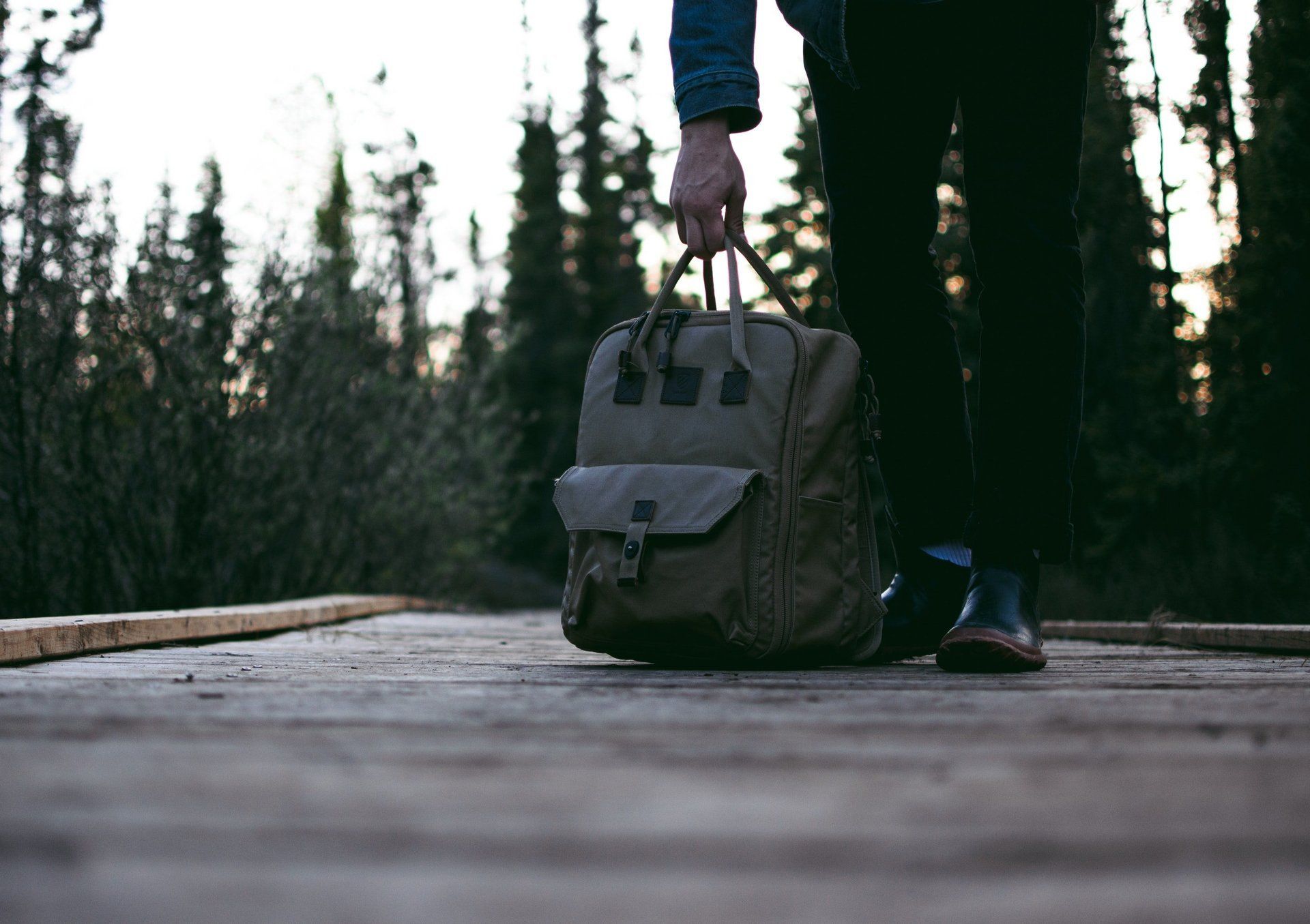 Person in blauer Jacke mit Rucksack auf einem Holzsteg im Wald in der Abenddämmerung
