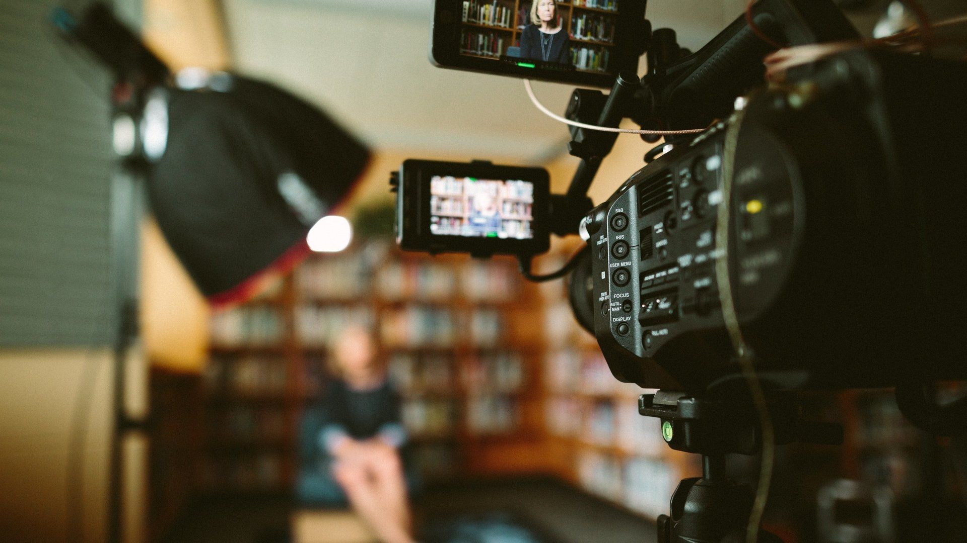 A video camera is recording a man sitting in front of a bookshelf.
