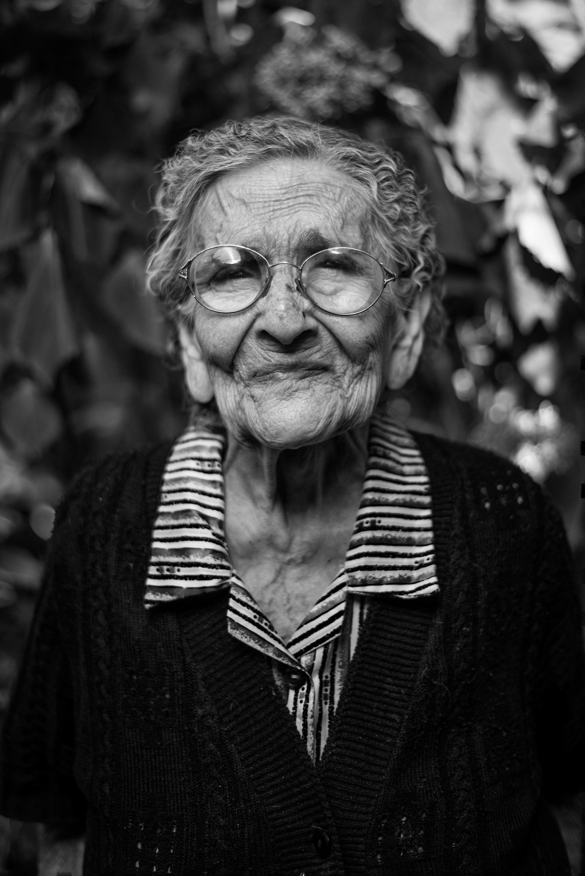 Elderly woman in glasses smiles at the camera, wearing a striped shirt and cardigan. Black and white photo taken outdoors.