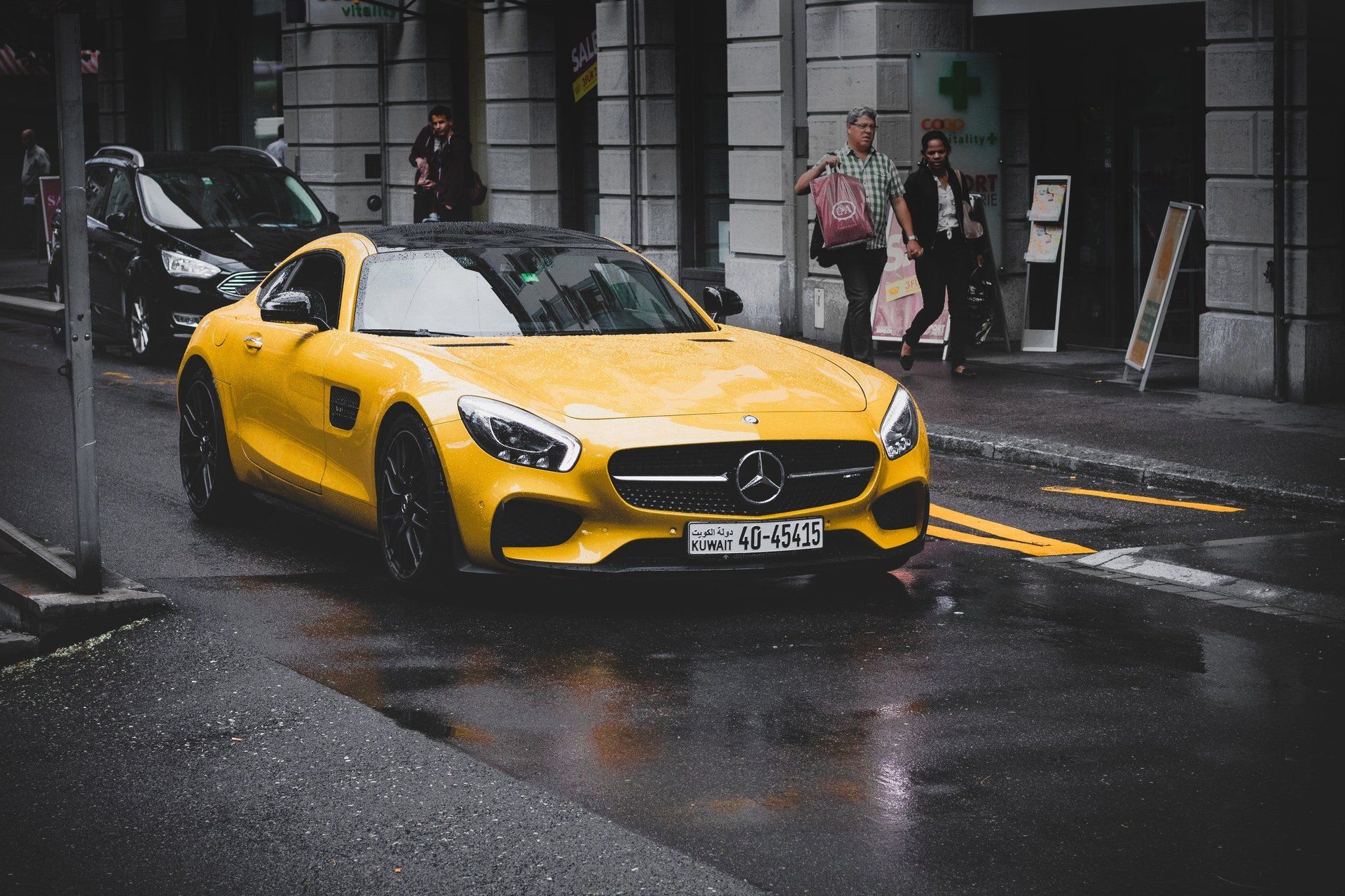 Yellow Mercedes-AMG GT sports car on a wet street, with people and buildings in the background.