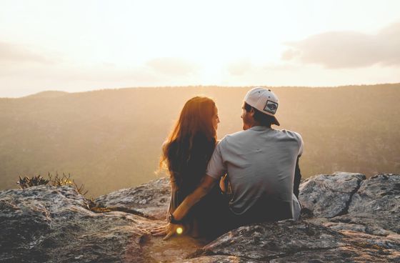 A man and a woman are sitting on top of a rock looking at the sunset.