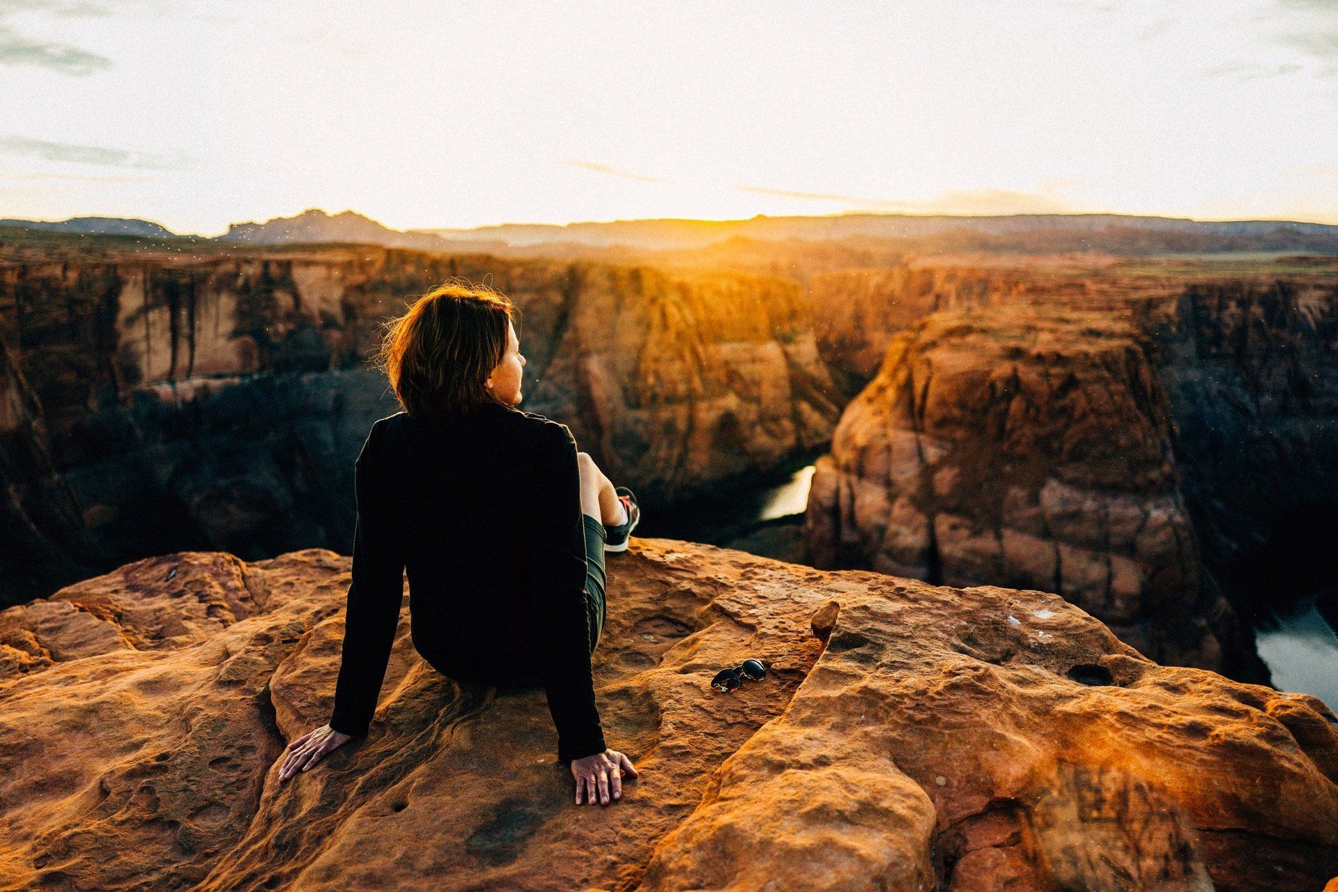 Person sitting on a rocky cliff at sunset, overlooking a canyon with warm golden light.