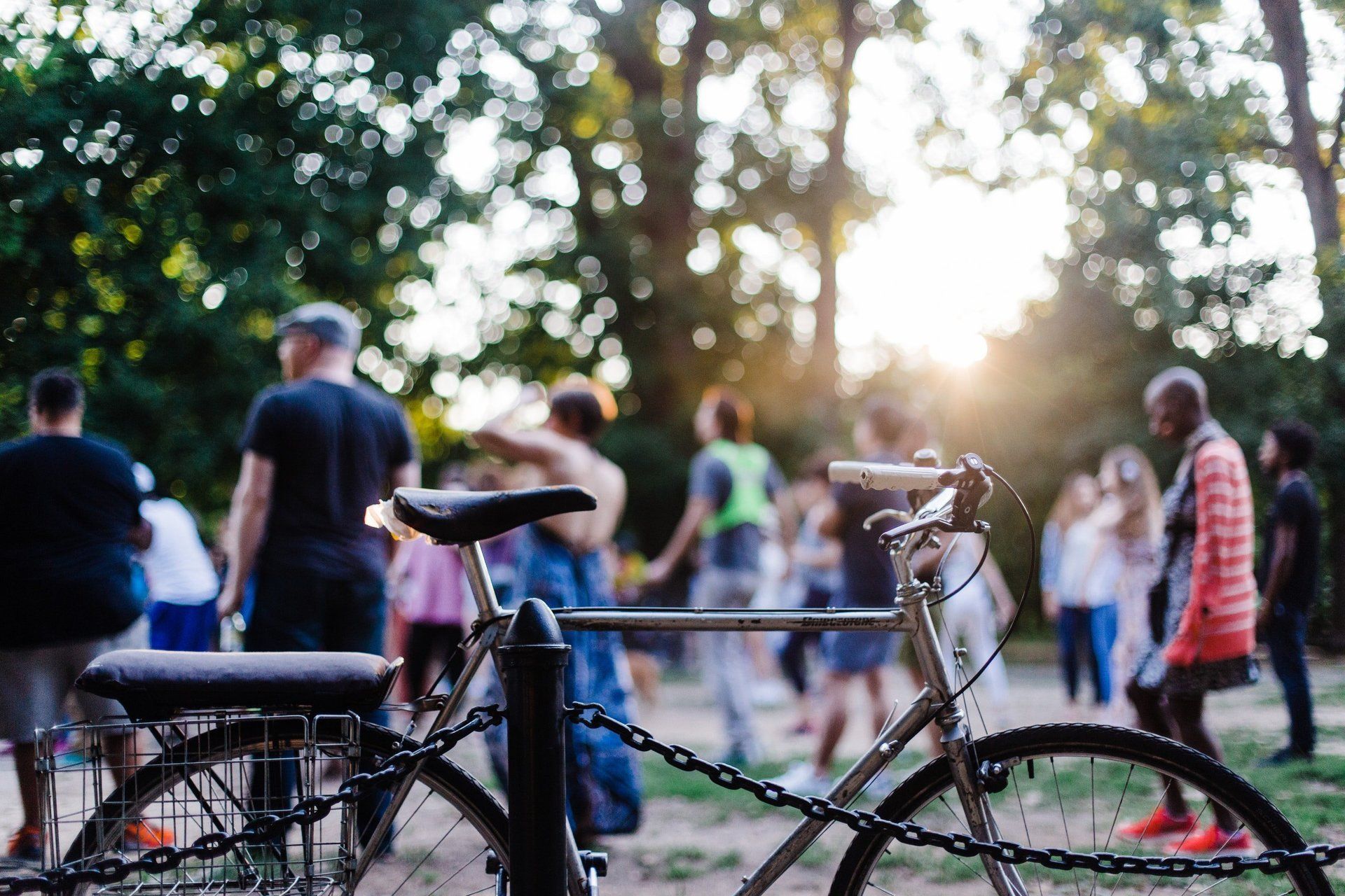 In primo piano, una bicicletta incatenata a un palo, mentre un gruppo di persone sfocate si riunisce in un parco al tramonto.