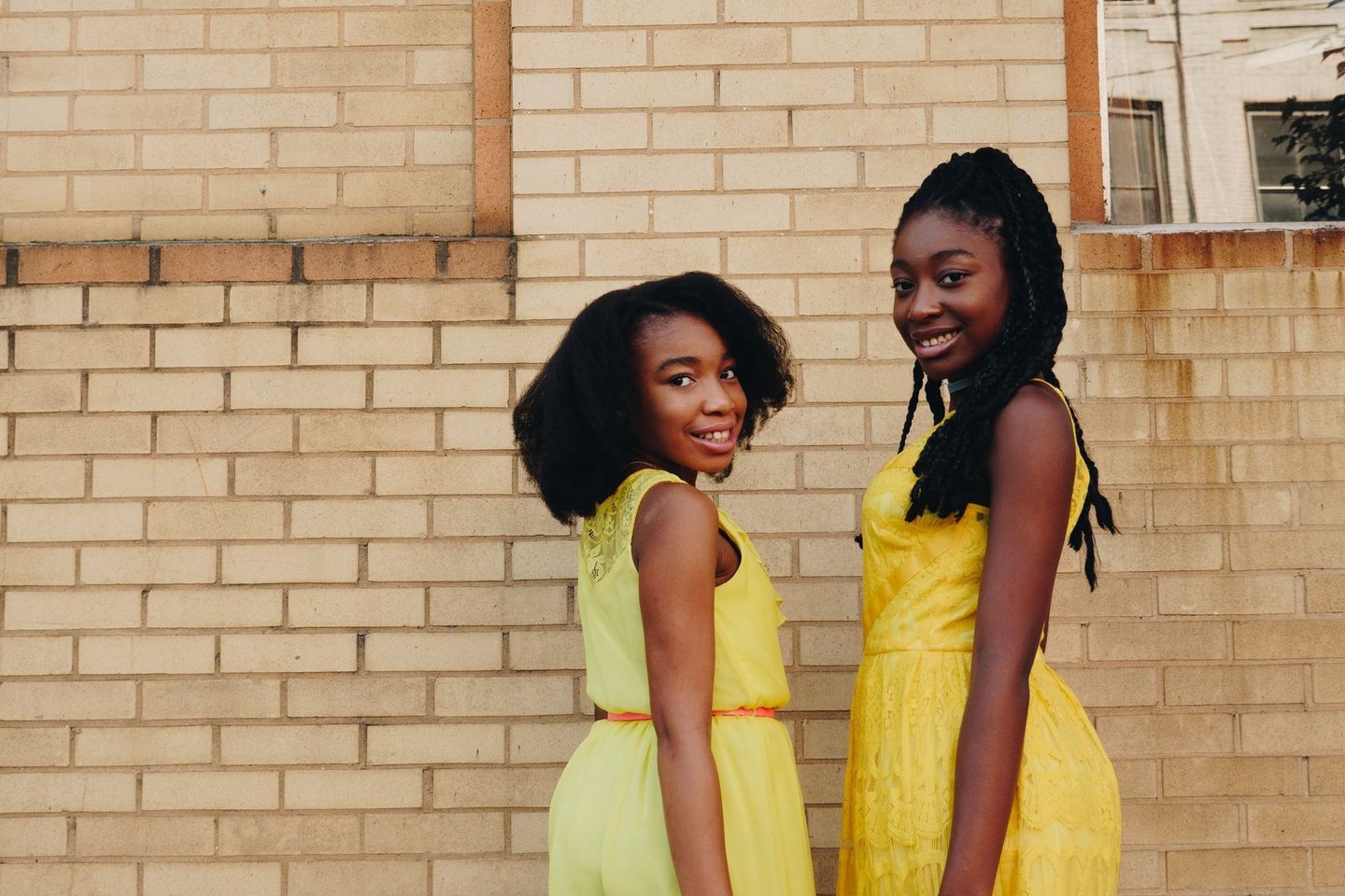 Two girls in yellow dresses are standing next to each other in front of a brick wall.