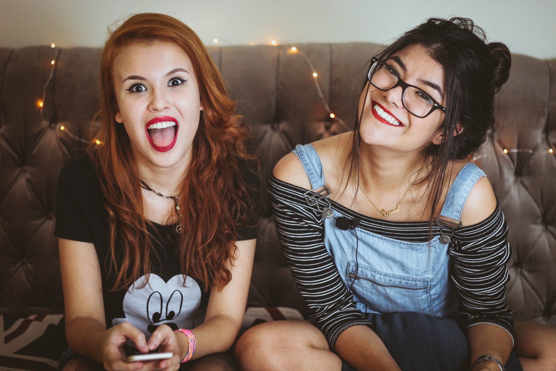 Two smiling friends sit on a couch, one holding a phone and making a playful face.