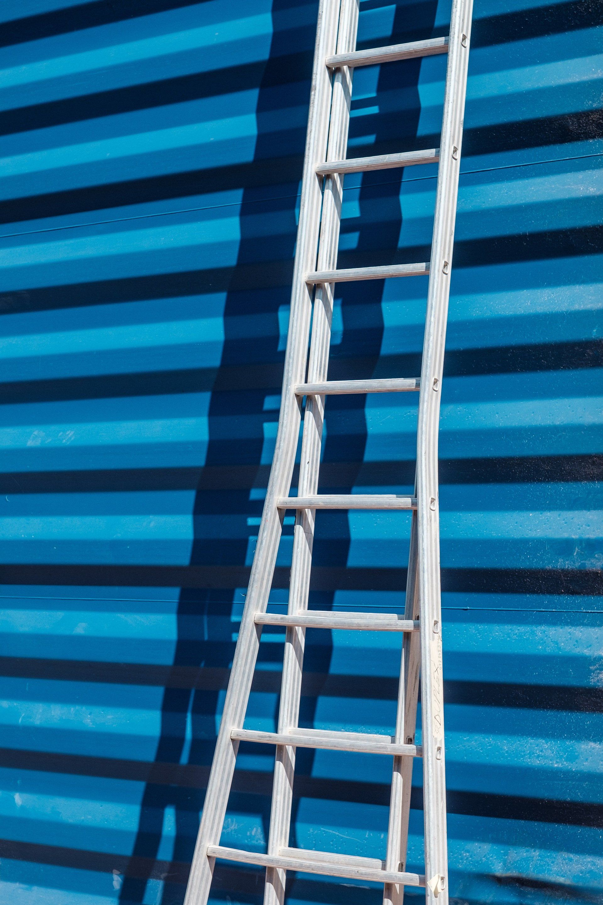 Silver ladder leaning against a blue corrugated wall, casting a shadow.
