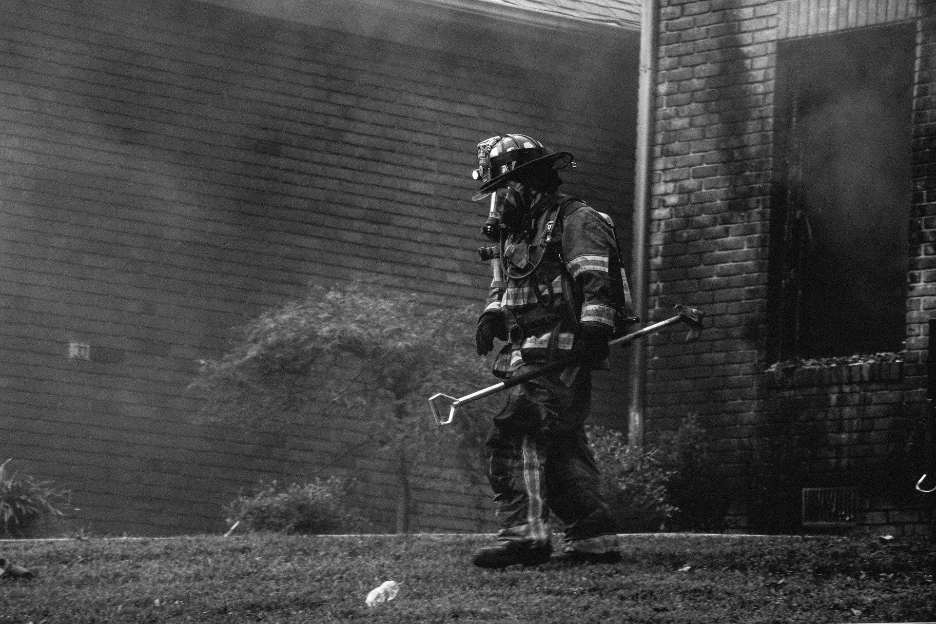 Ein Feuerwehrmann geht auf einer Wiese in der Nähe eines brennenden Gebäudes und hält ein Werkzeug in der Hand.