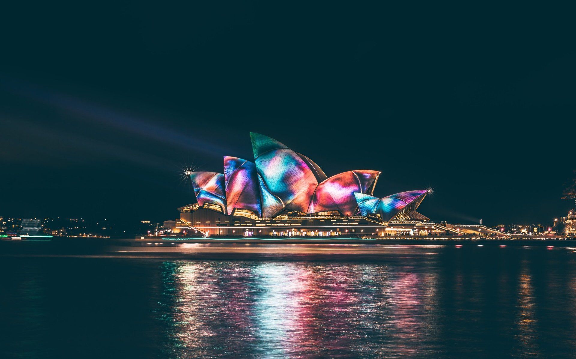 The sydney opera house is lit up at night and reflected in the water.