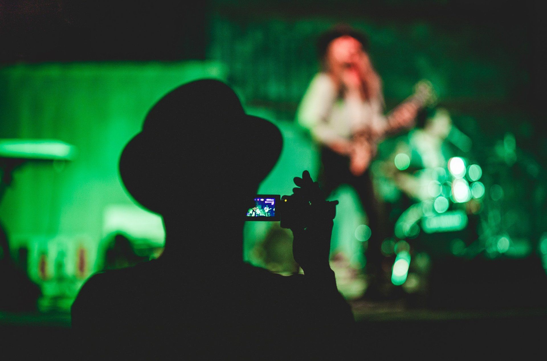 Silhouetted audience member filming a green-lit concert with a singer on stage in the background