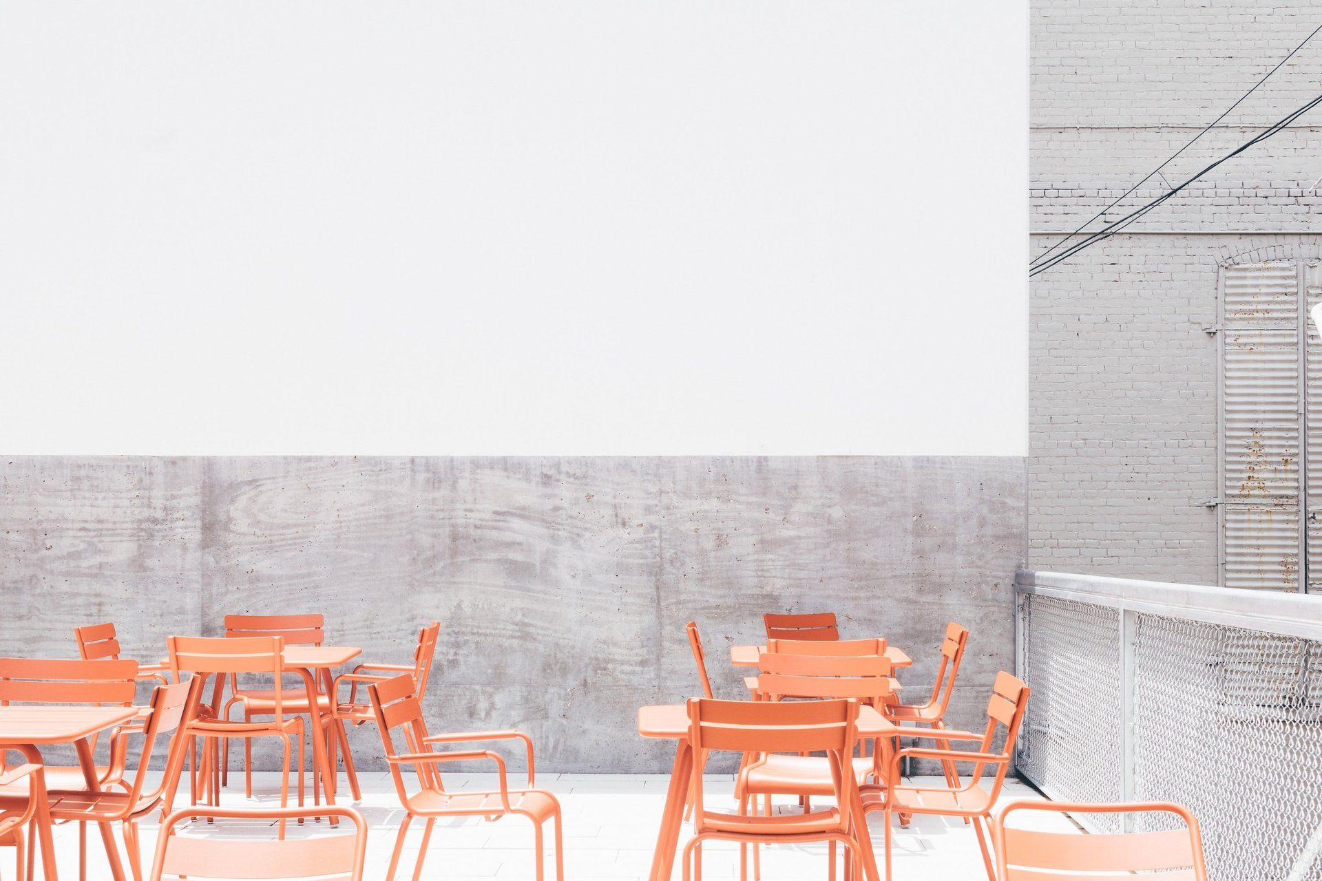 Una terrazza con sedie e tavoli arancioni, addossata a un muro grigio chiaro e a una recinzione dalla texture particolare.