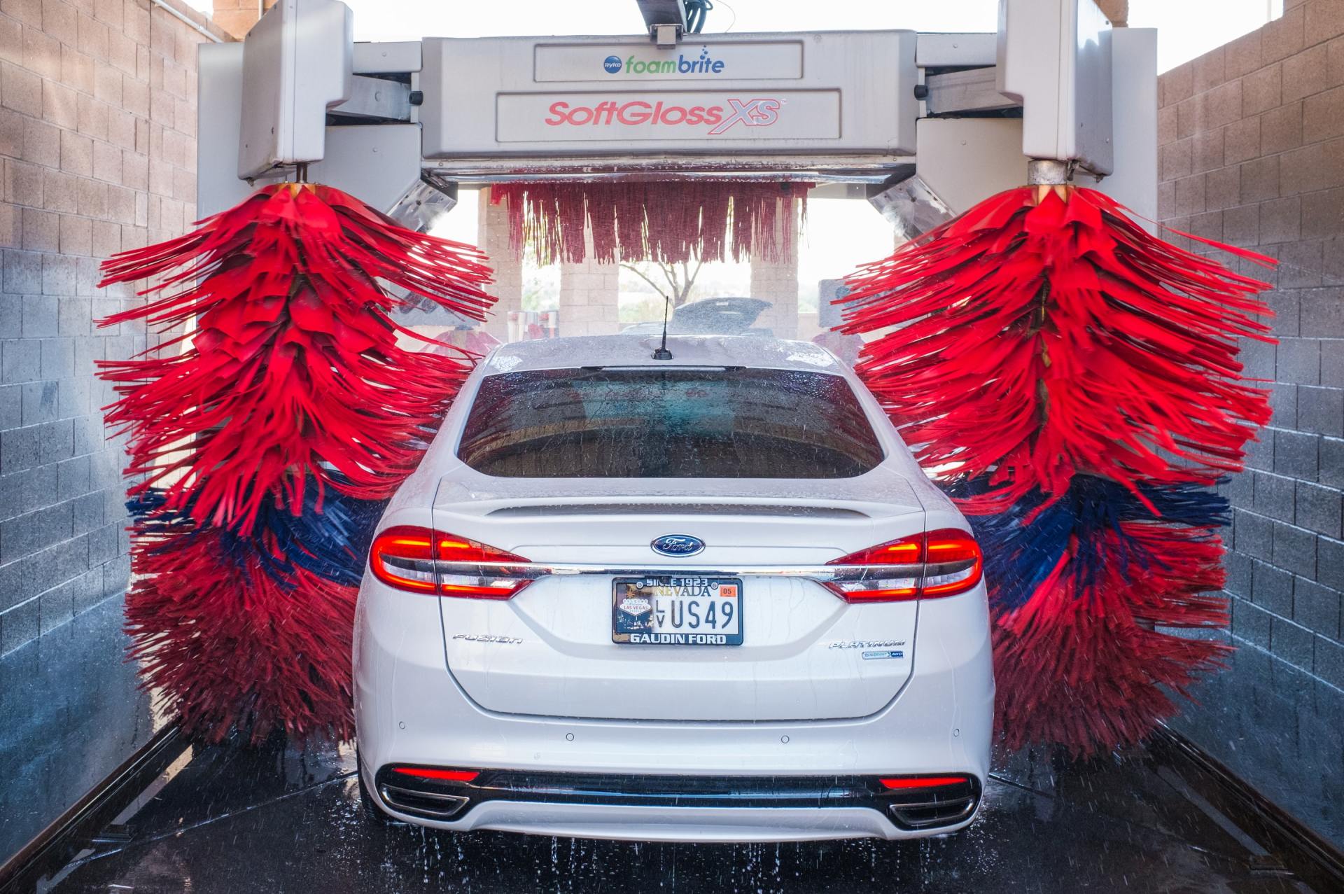 A white car is going through a car wash with red brushes.