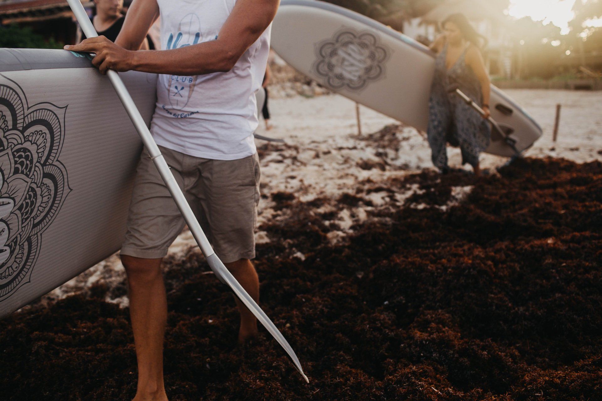 A man is carrying a surfboard and a paddle on the beach.