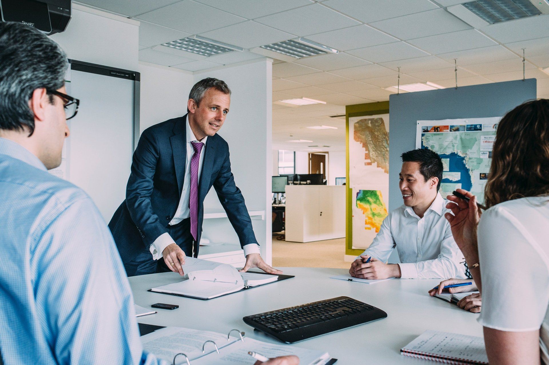 Business meeting: Man in suit speaks to colleagues seated around a table. Open office setting.