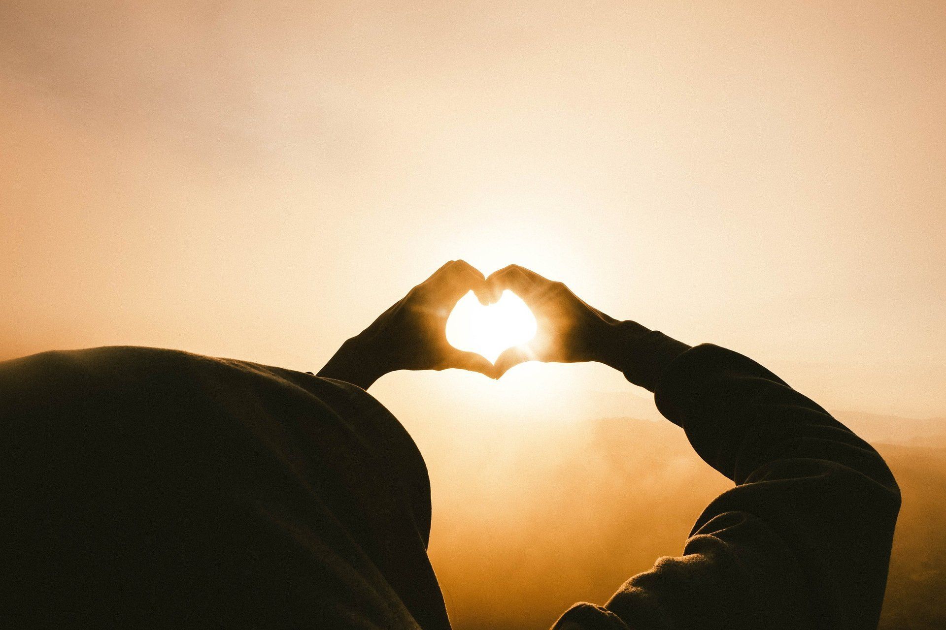 A person is making a heart shape with their hands in front of the sun.