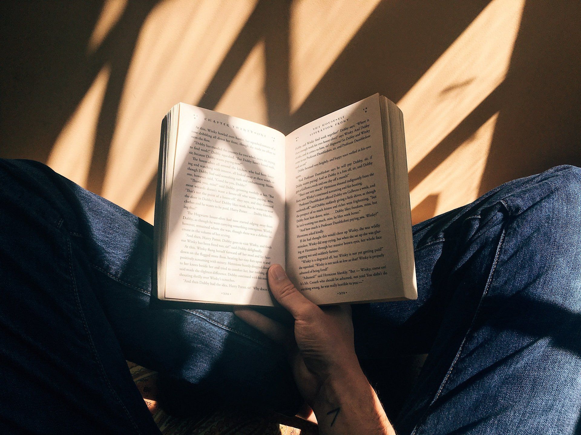 Open book in lap, sunlight casting shadow, denim jeans.