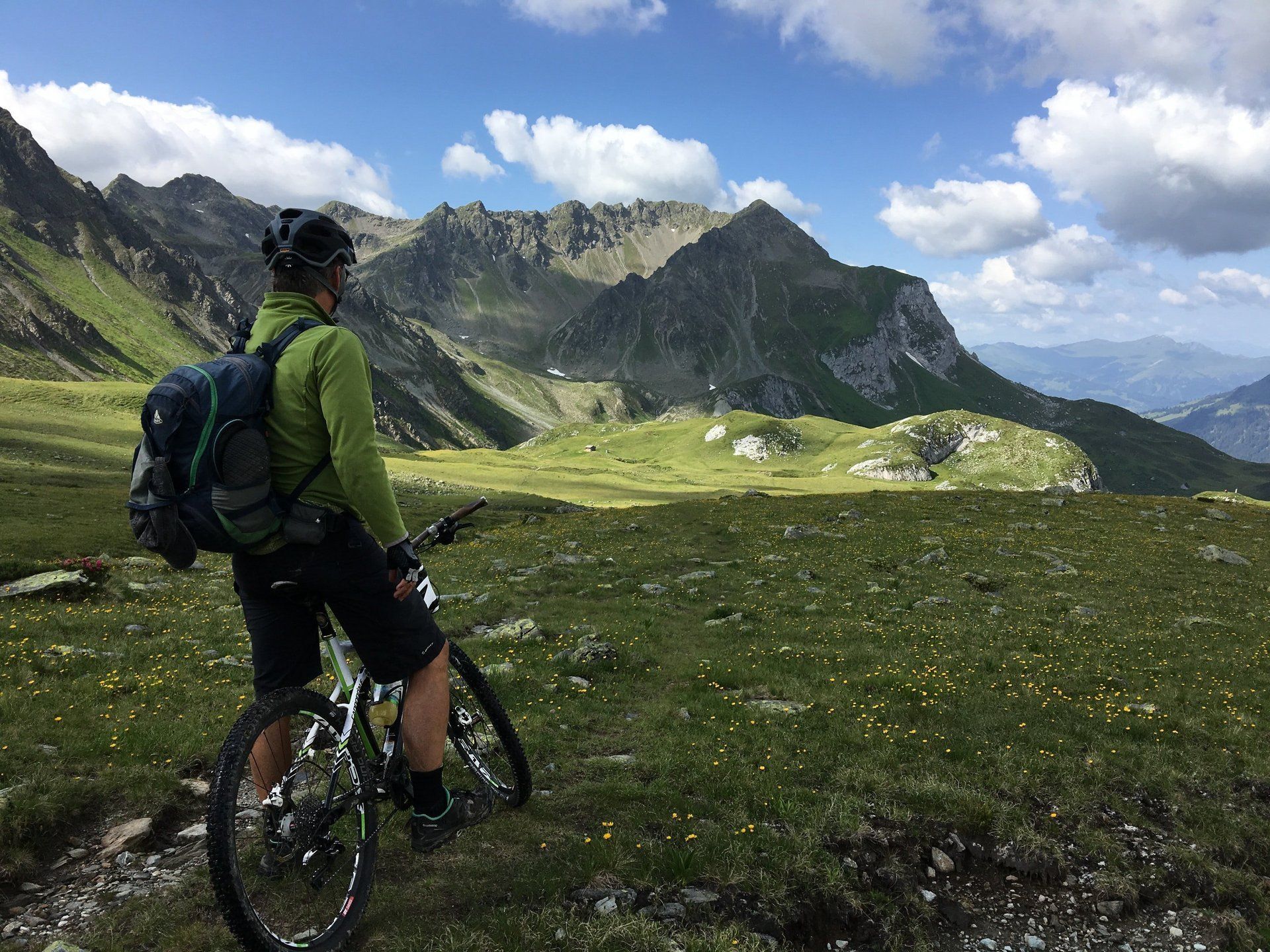 Radfahrer mit Rucksack auf einem Bergpfad mit Blick auf grüne Alpengipfel unter blauem Himmel