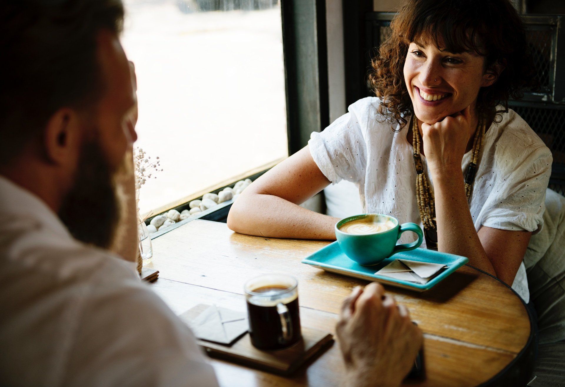 Two people sit at a wooden table in a café, drinking coffee and smiling while engaged in conversation.