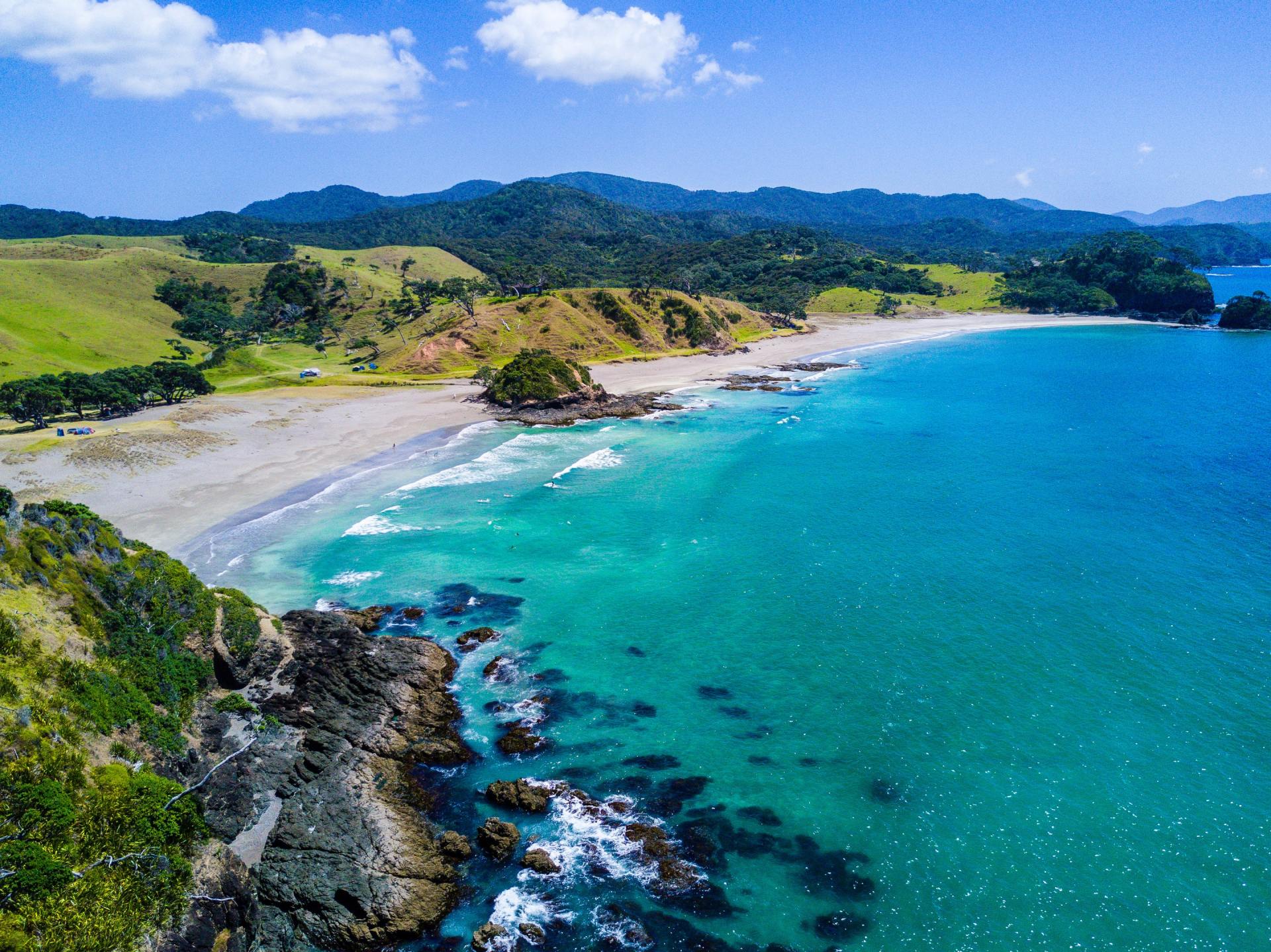 Tropical coastline with turquoise water, sandy beach, rocky shore, and green hills under a blue sky