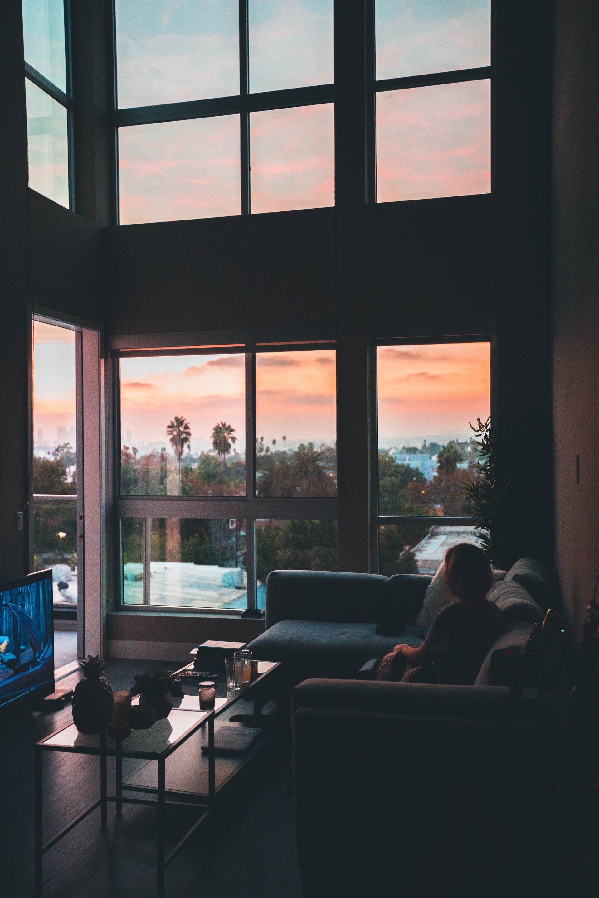 Living room with large windows overlooking a sunset, person on couch, television.