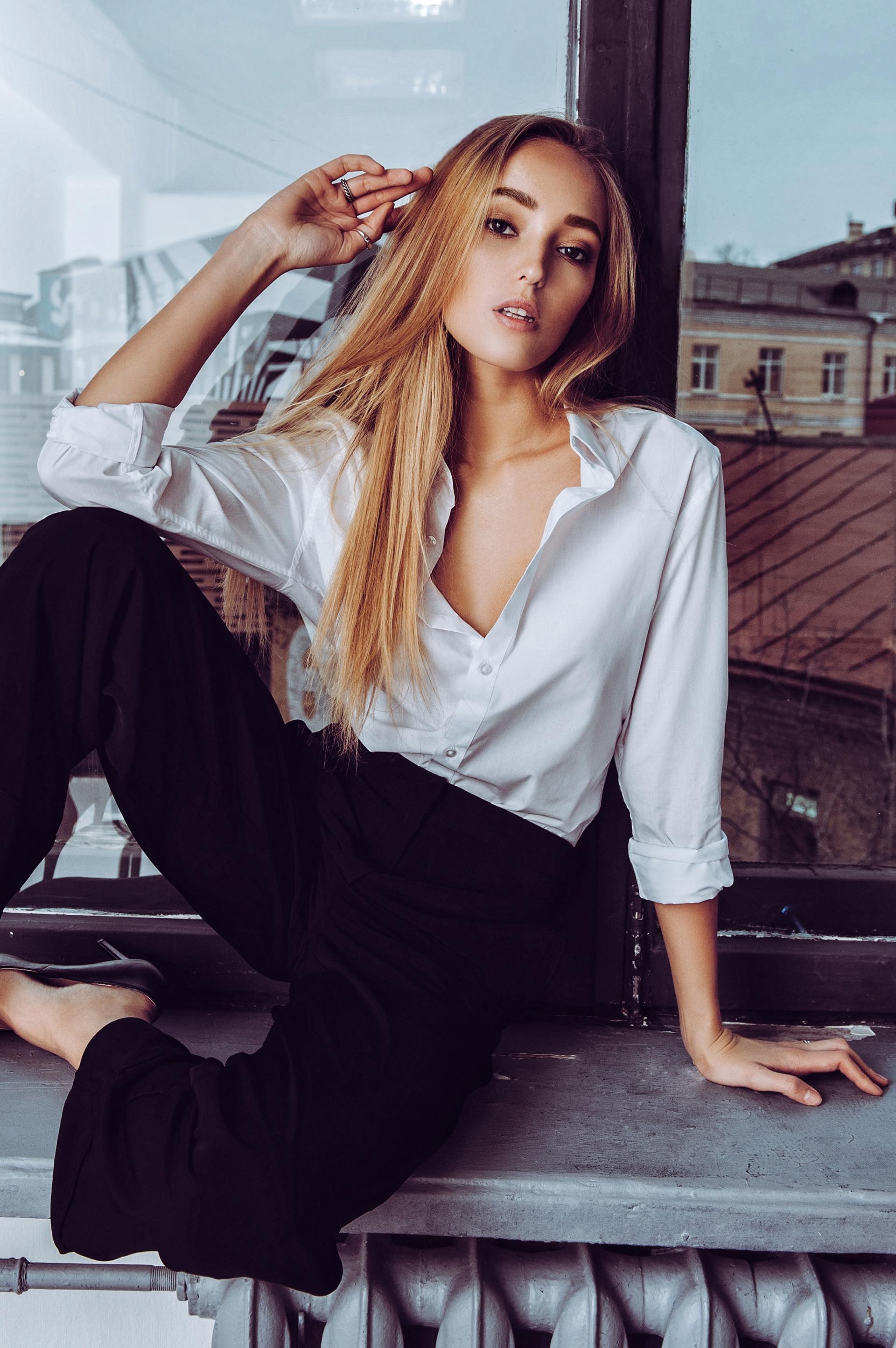 A woman in a white shirt and black pants is sitting on a window sill.