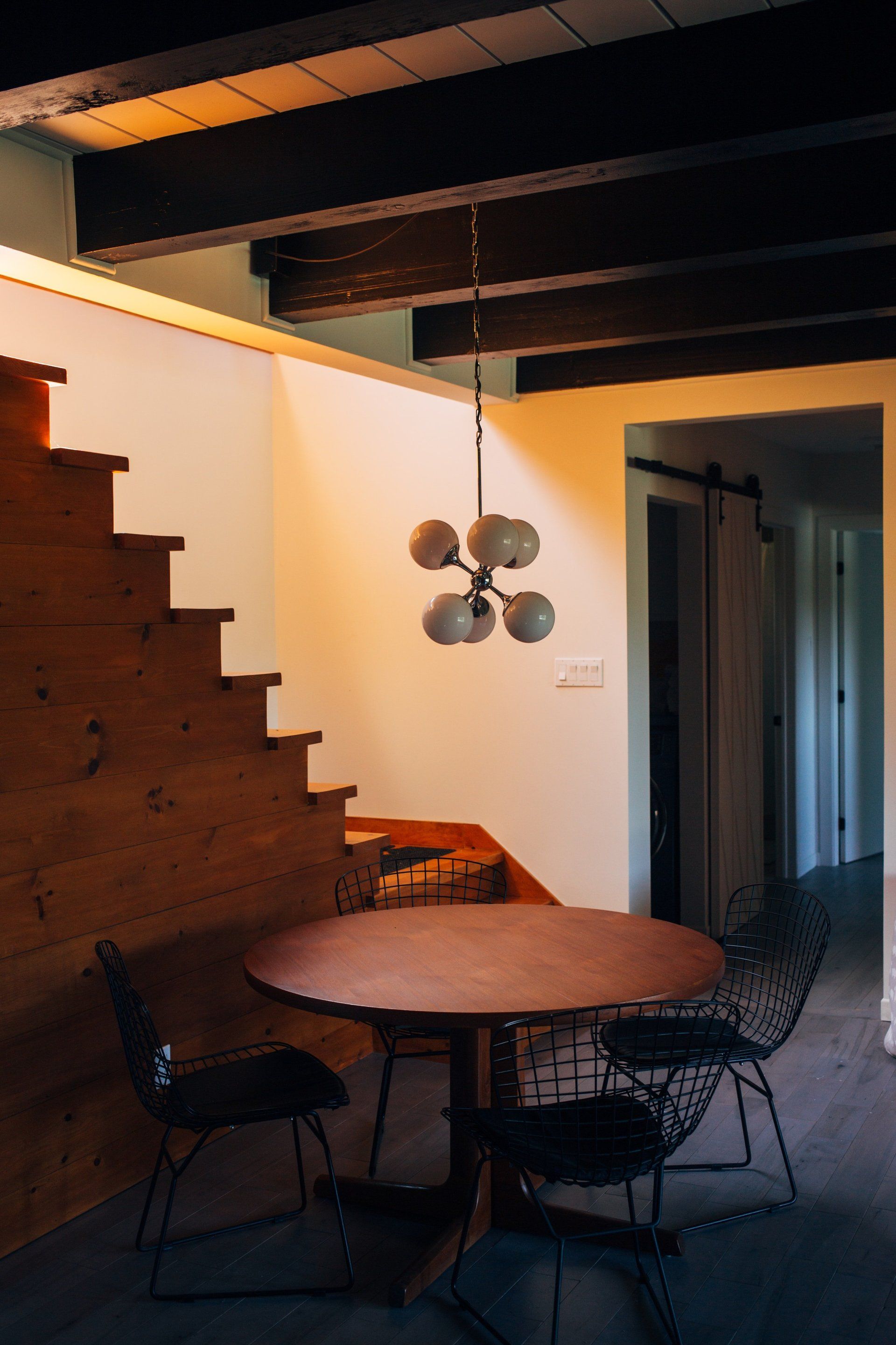 Small dining area with round wooden table, black chairs, staircase, and wall-mounted fan in warm lighting