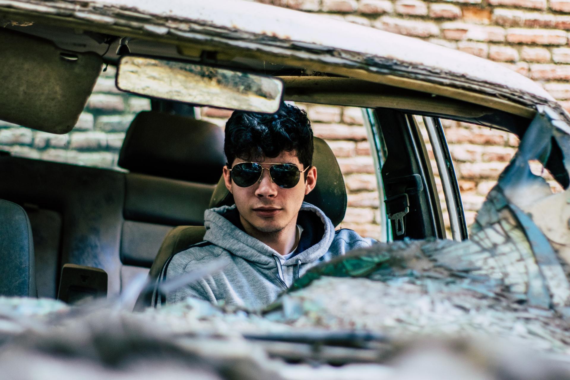 Man in sunglasses sits in a wrecked car, staring forward. Windshield is shattered.