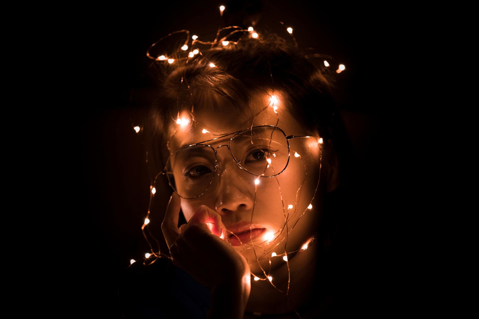Person with string lights wrapped around their face, in a dark setting, looking at the camera.