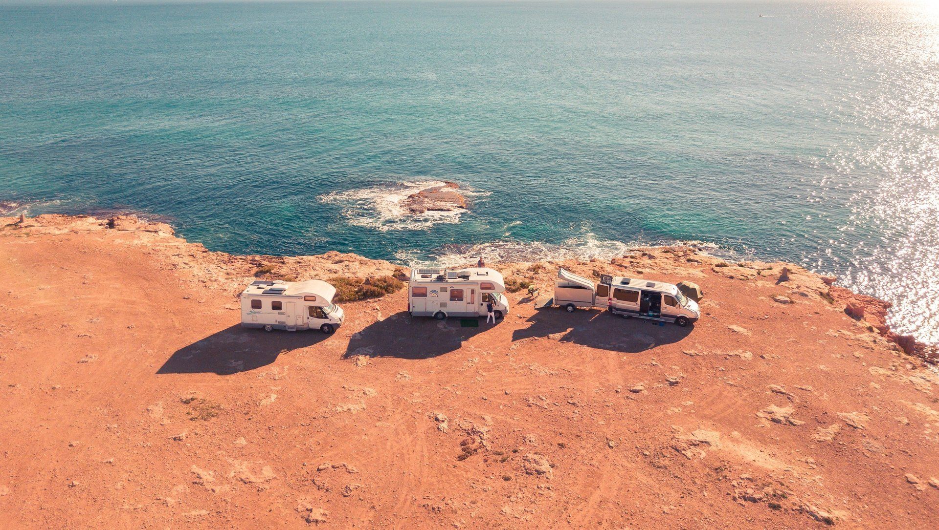 Three campers are parked on a rocky cliff overlooking the ocean under a clear sky.