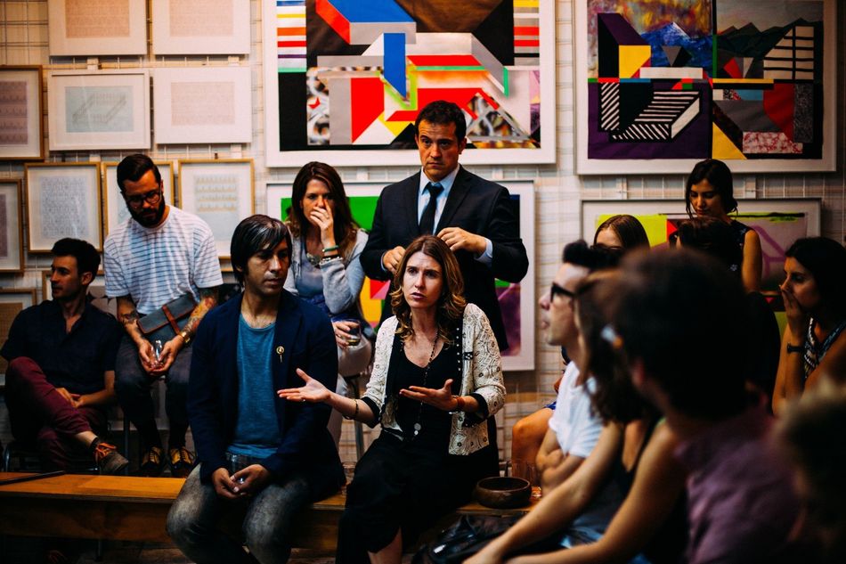 People gathered at an art gallery, listening to a woman speaking. Paintings on the wall.