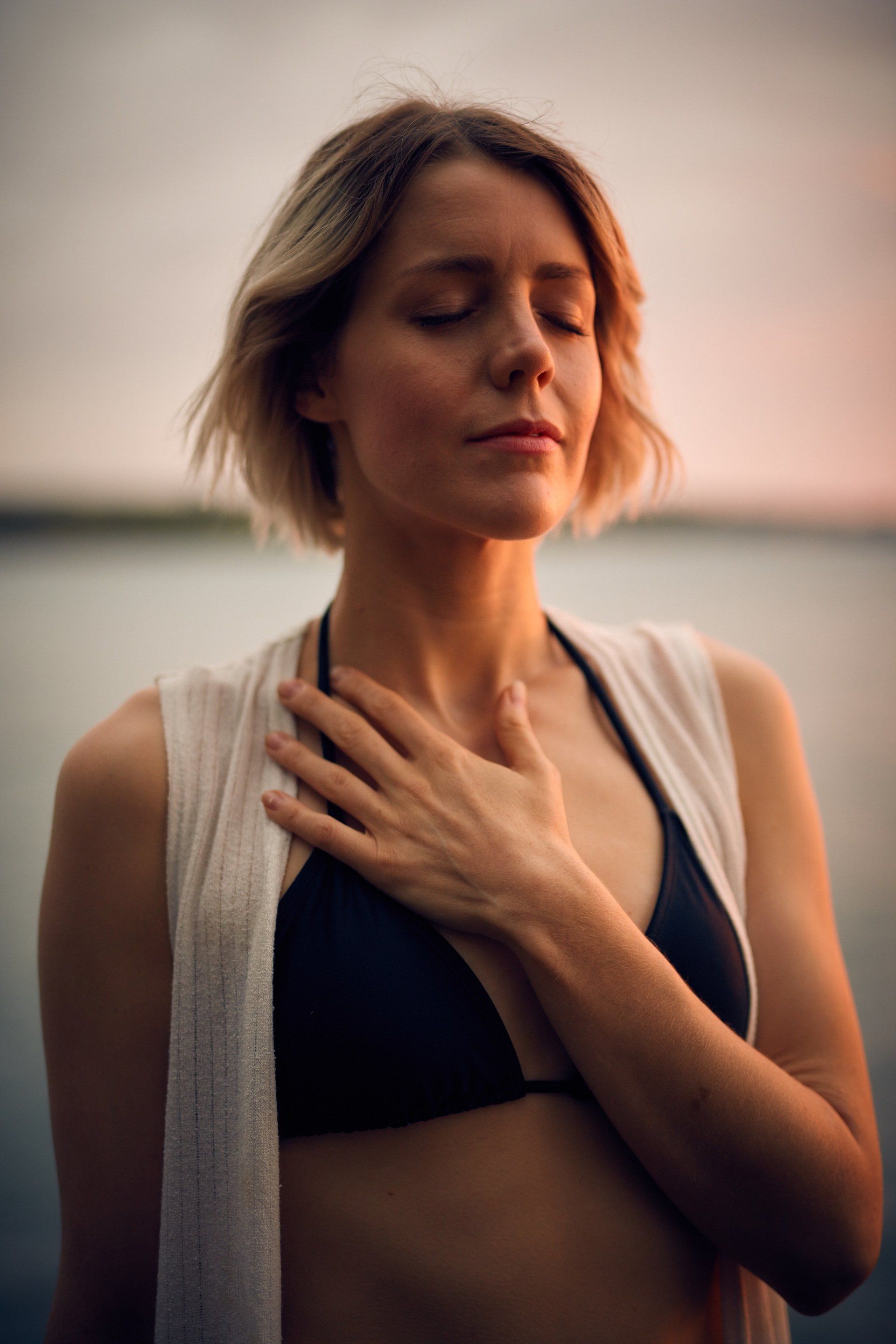 Woman with eyes closed, hand on chest, wearing a bikini top and cardigan, near water with a sunset in the background.