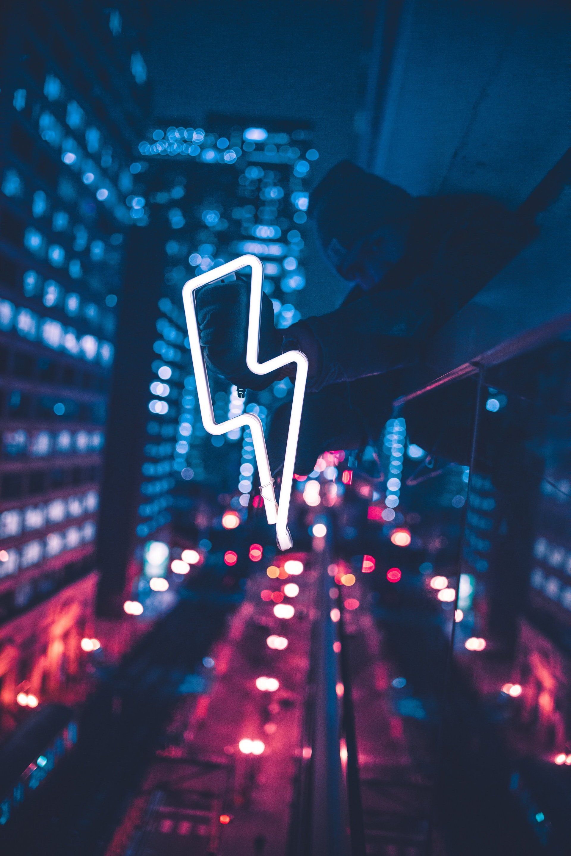 A neon lightning bolt is hanging from a railing in the middle of a city at night.