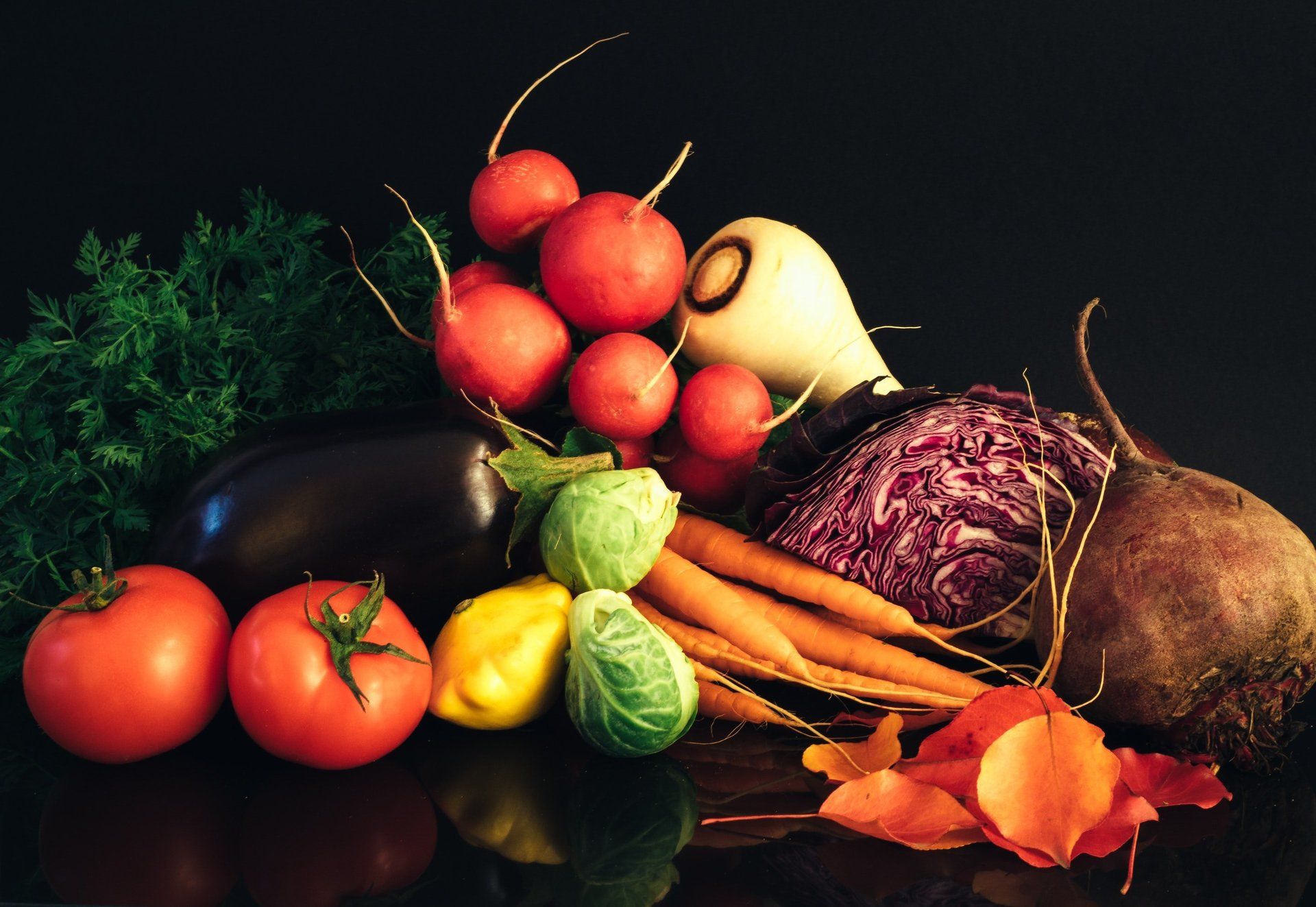 Still life of various colorful vegetables, including radishes, tomatoes, and carrots, arranged on a dark surface.