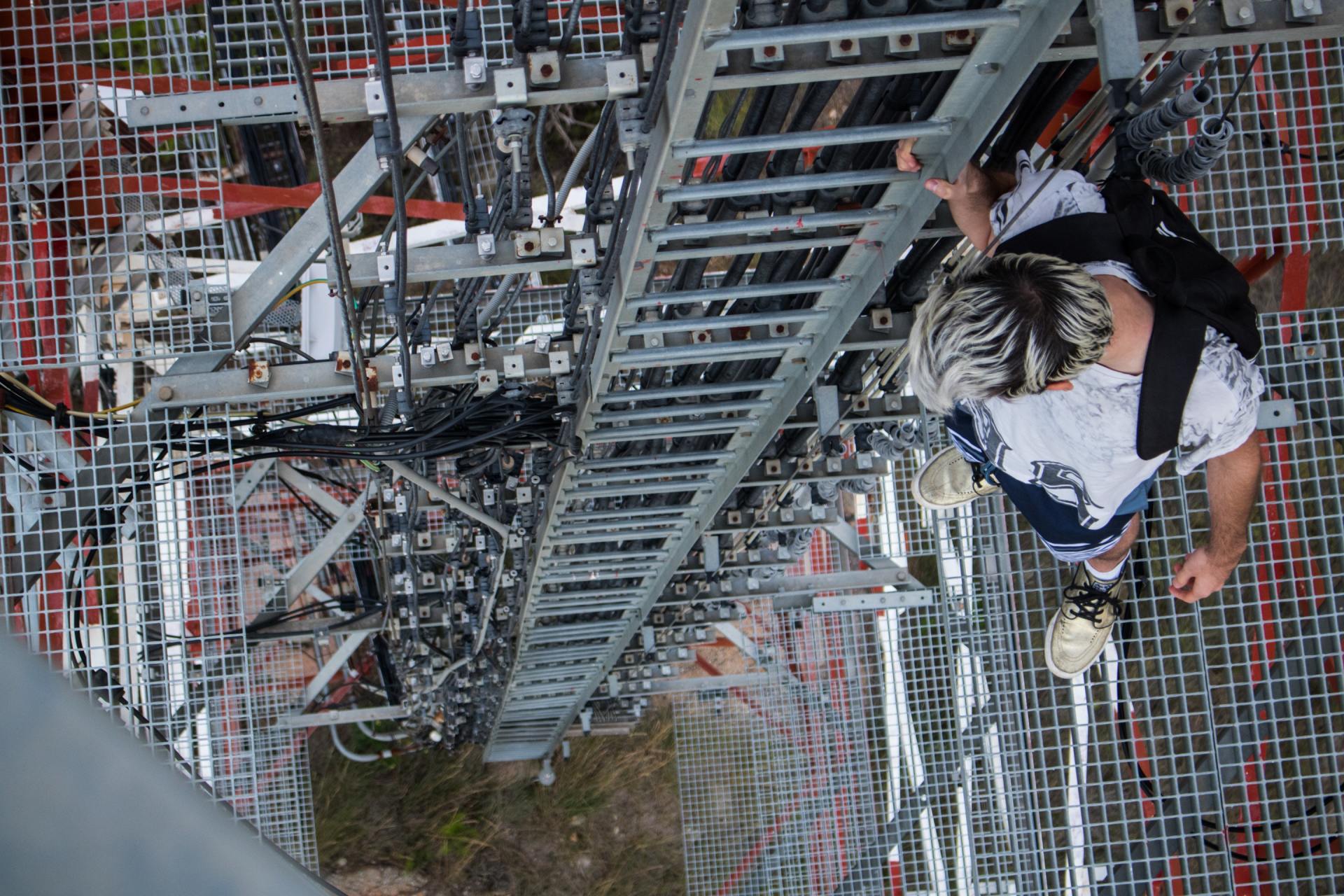 Person on a metal platform, looking down at a ladder on a tall structure, possibly a communication tower.