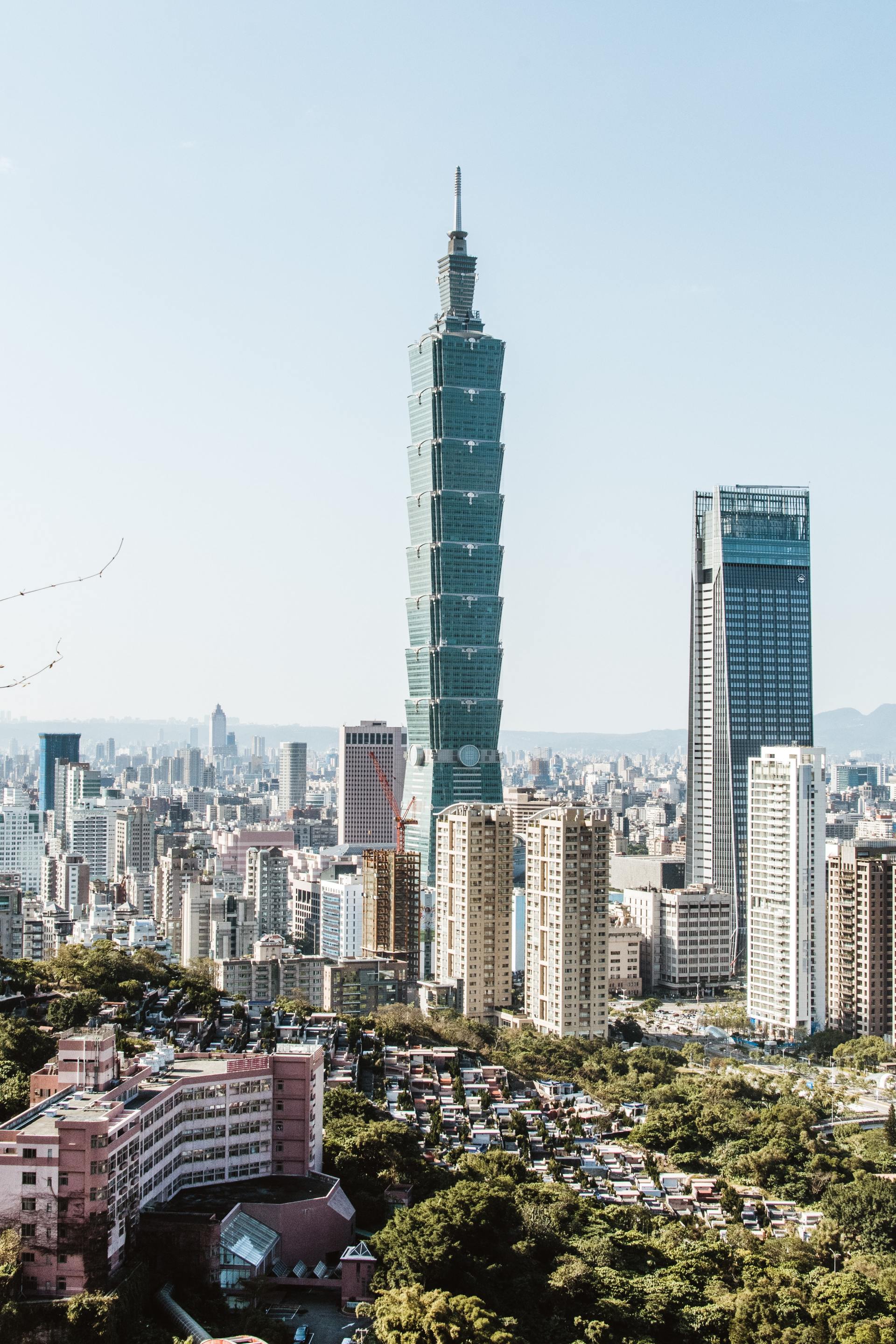 Taipei city skyline with prominent green-tinted Taipei 101 tower. Other buildings and greenery visible on a sunny day.